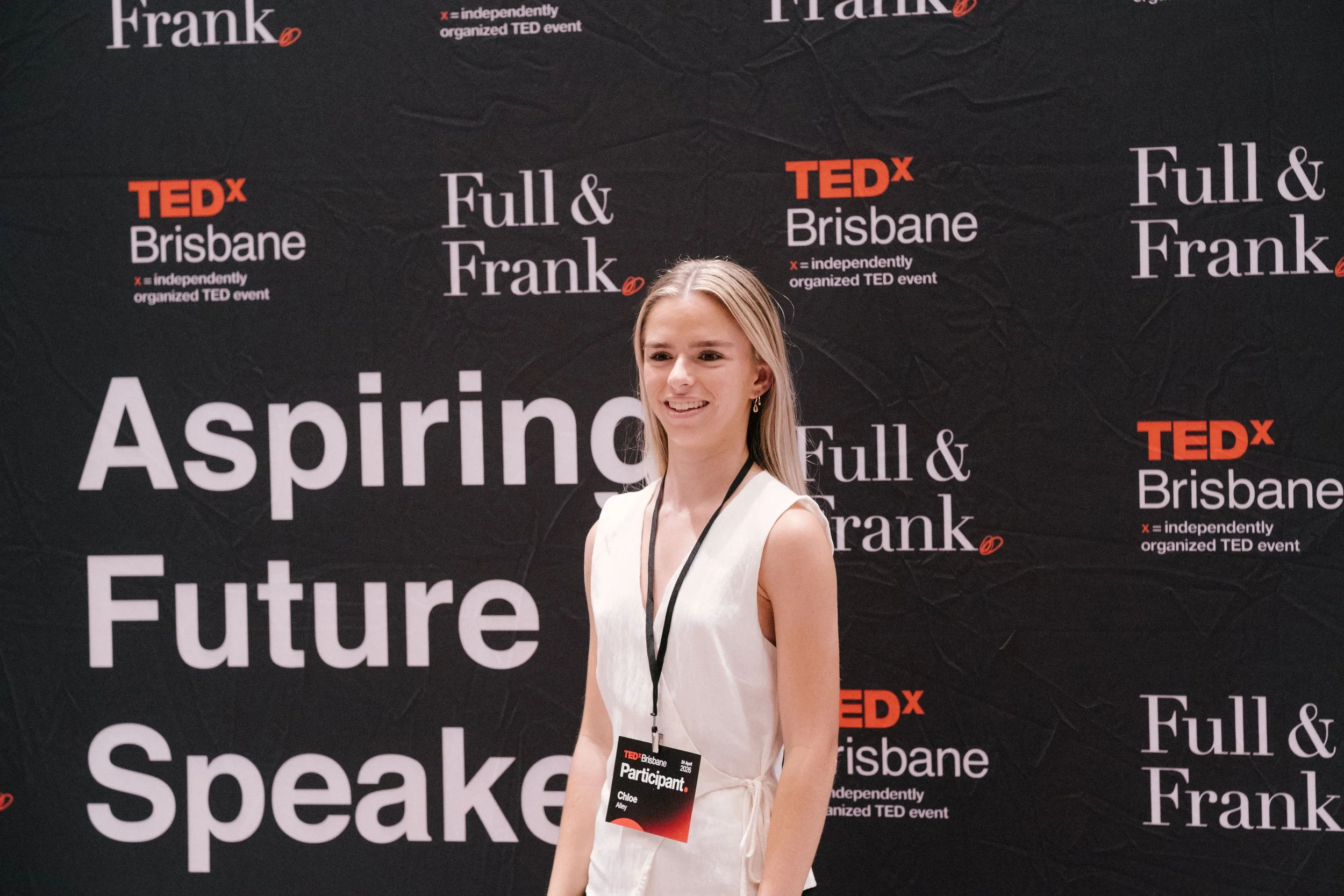 Young woman standing in front of a black TEDx Brisbane backdrop with the words "Full & Frank" repeated. She is wearing a light-colored dress, earrings, and a lanyard with a TEDx participant badge that reads "Chloe".