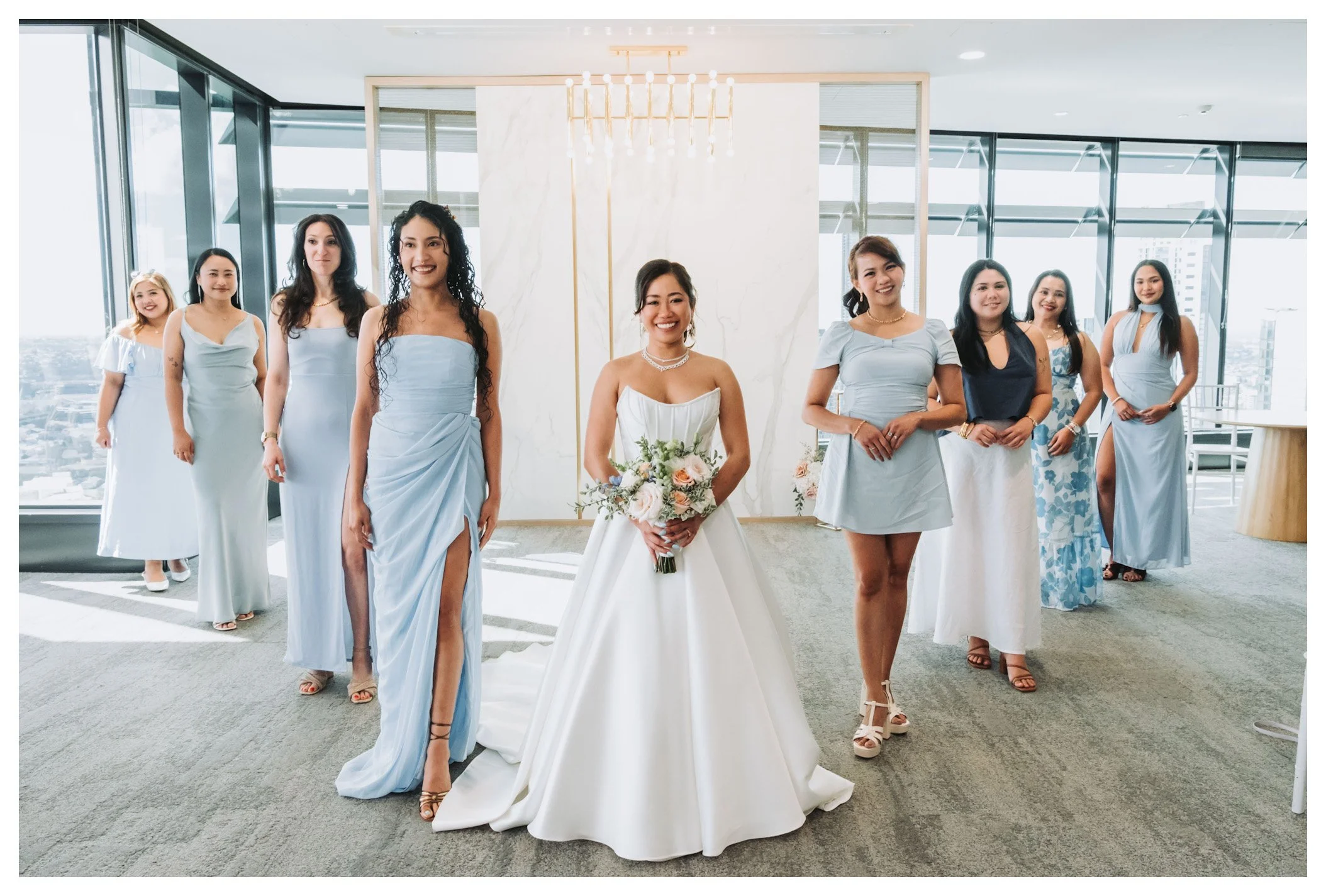 A bride in a strapless white wedding dress holding a bouquet, surrounded by eight women in light blue and blue dresses, posing on a high-rise building with city skyline view through large windows.