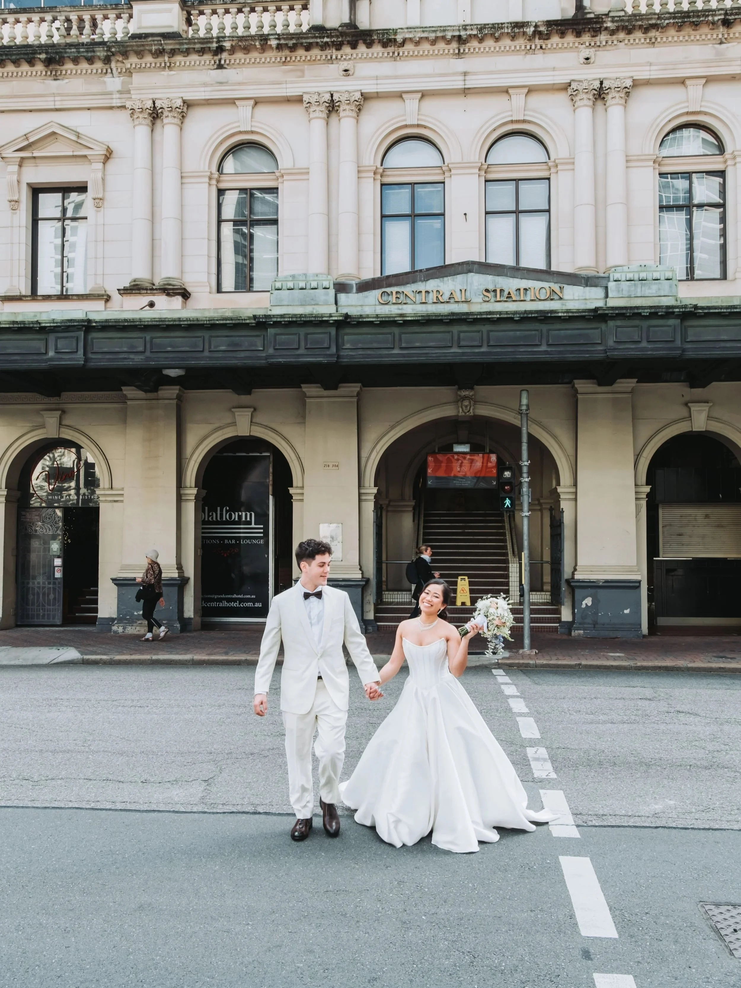Bride and groom walk hand-in-hand across a street in front of Central Station in elegant wedding attire.