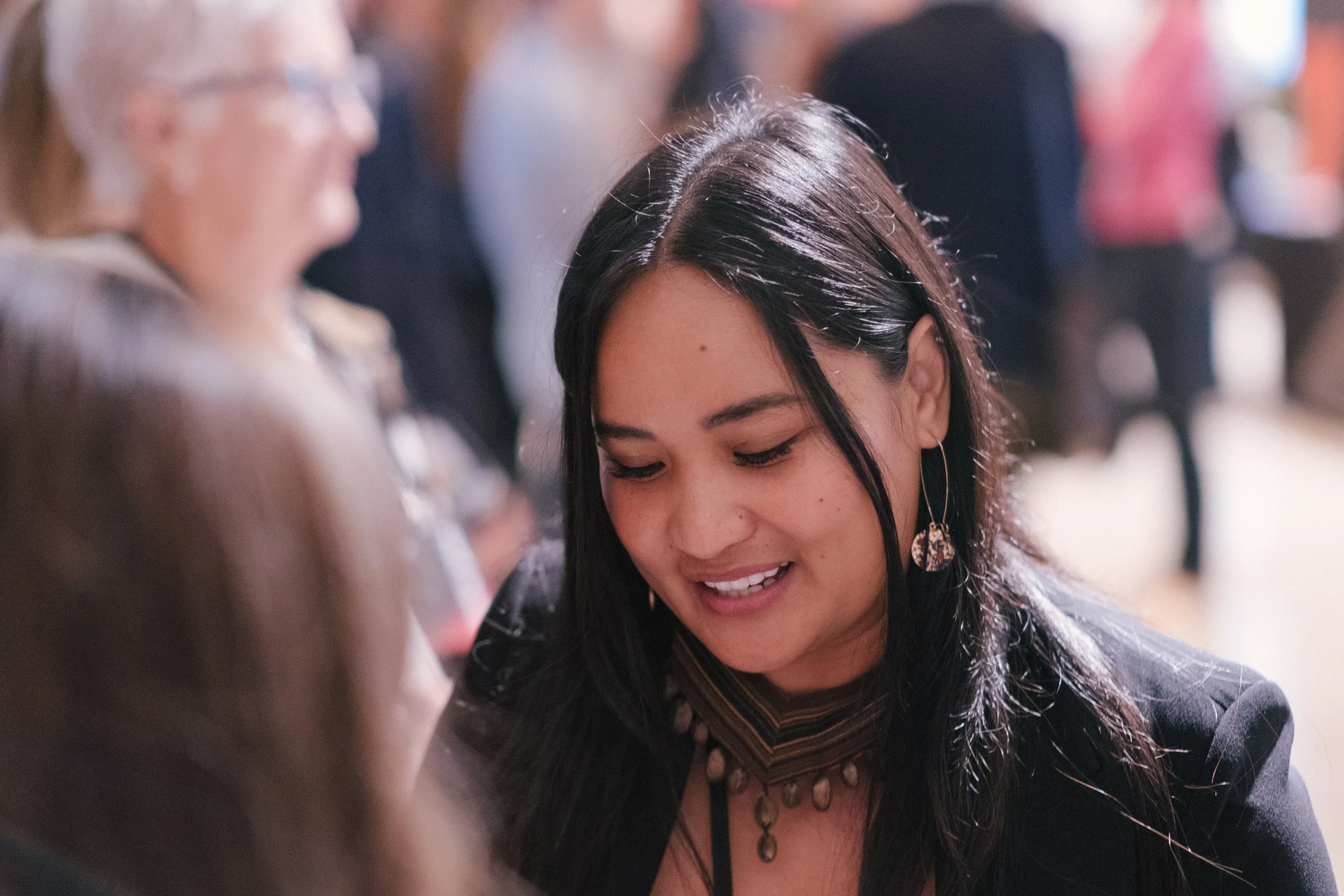 A woman with dark hair, earrings, and a necklace is smiling and looking down. She is having a conversation or sharing a moment with another person, whose face is blurred and who is on the left side of the image.