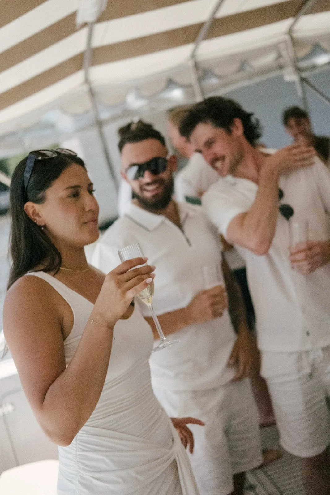 A woman in a white dress holding a champagne glass at a social gathering with people in white attire, some wearing sunglasses, smiling and talking under a tent.