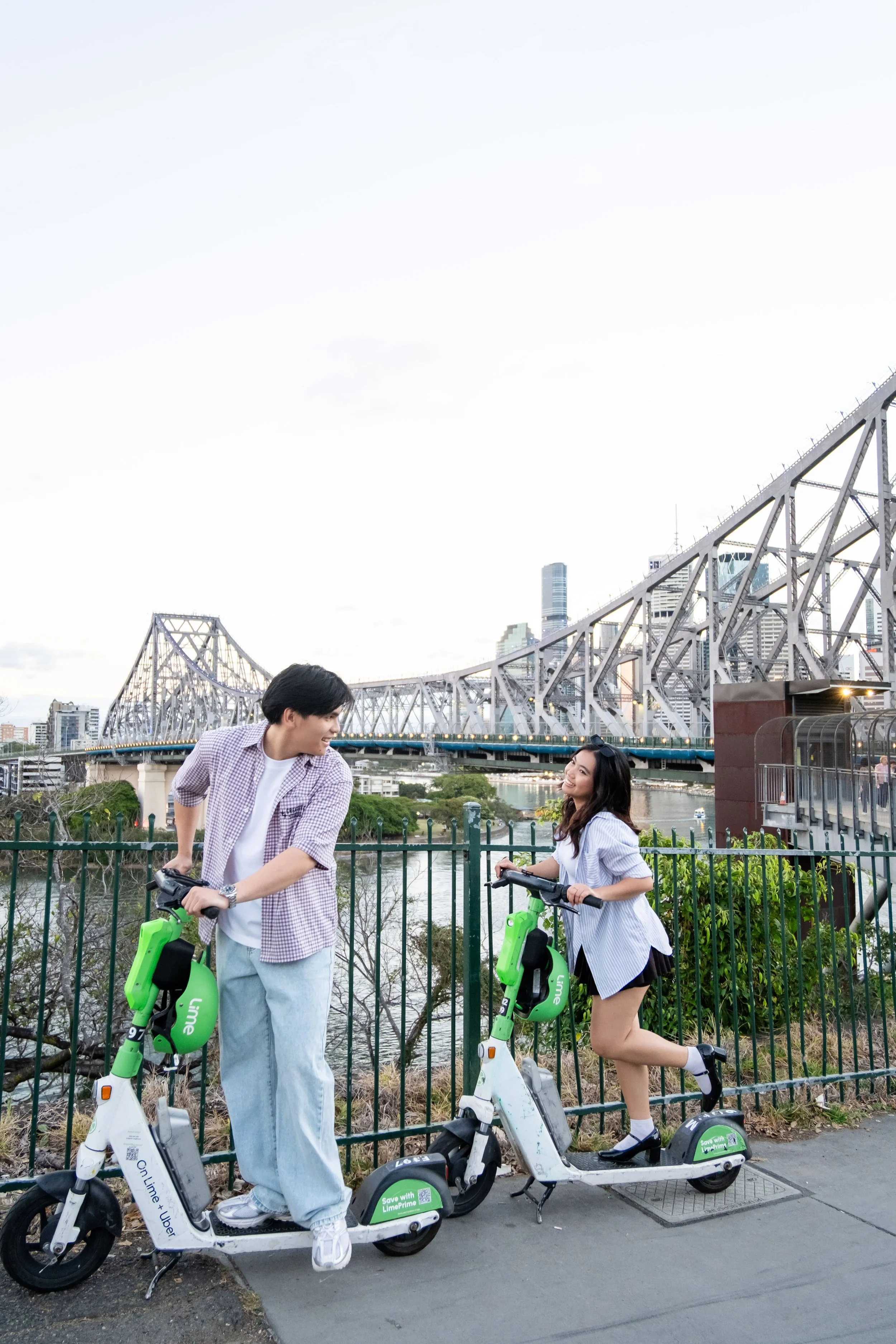 A young man and woman smiling and talking while standing on Lime scooters near a fence by a river, with a bridge and city skyline in the background.