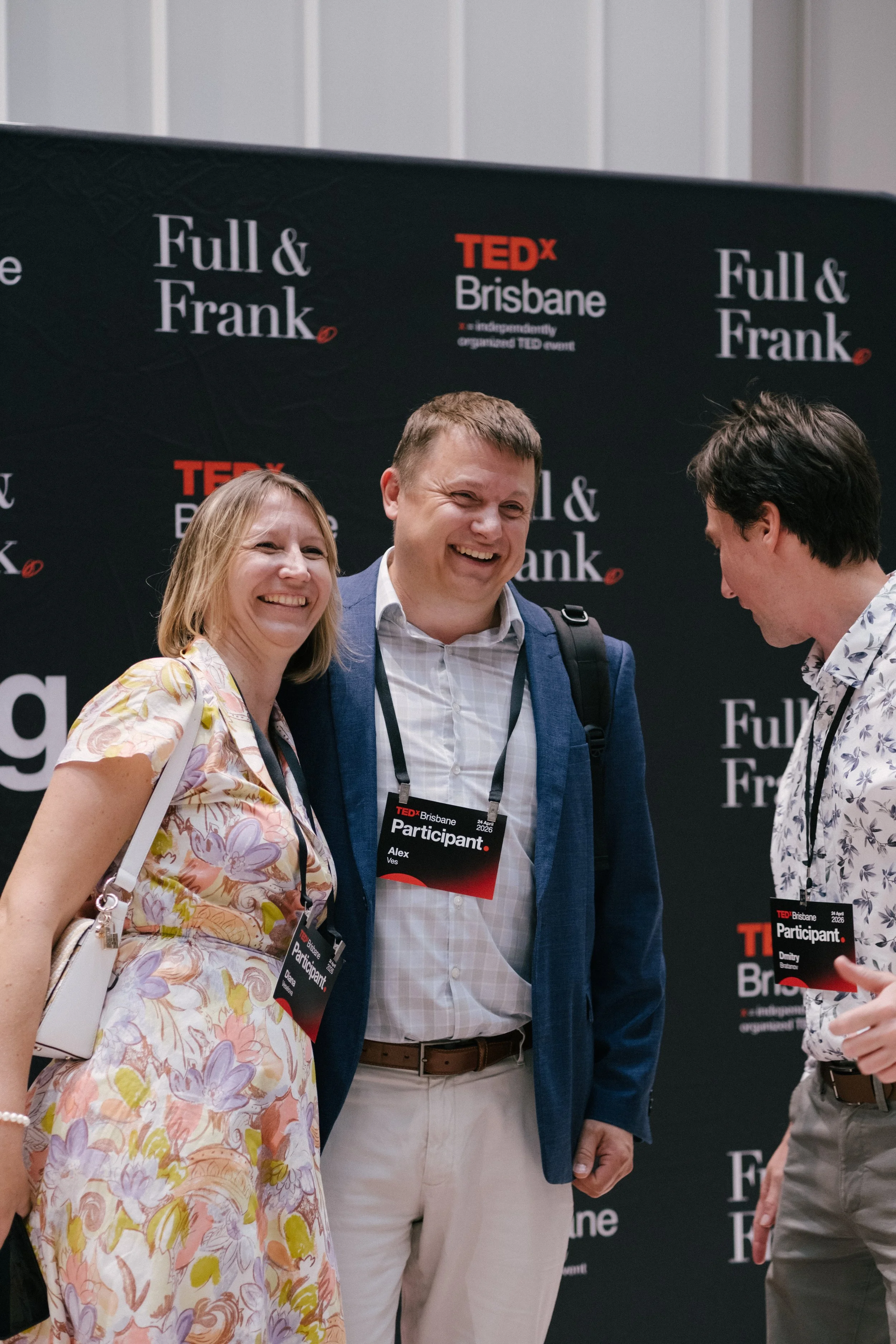 Three people at a TEDx Brisbane event, standing in front of a black backdrop with the TEDx Brisbane logo and event branding. The woman on the left is wearing a floral dress, has shoulder-length blonde hair, and is smiling broadly. The man in the midd