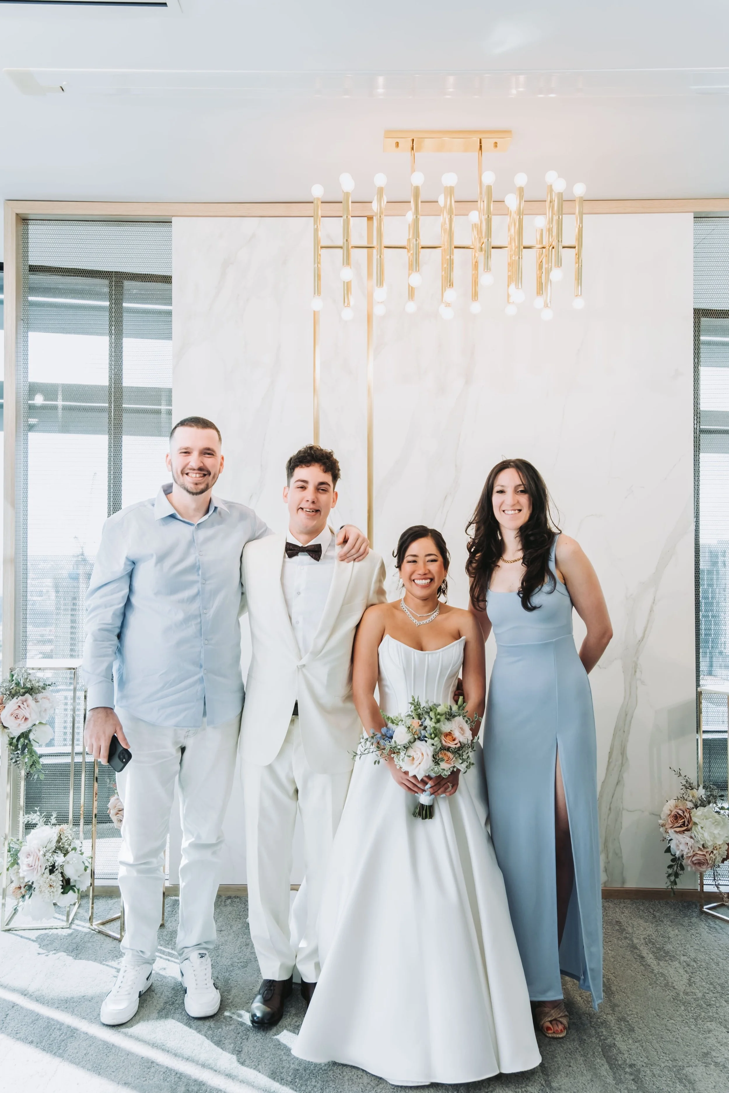 A group of five people dressed in formal attire, including a bride holding a bouquet, standing together indoors in front of a white marble wall and a modern chandelier.