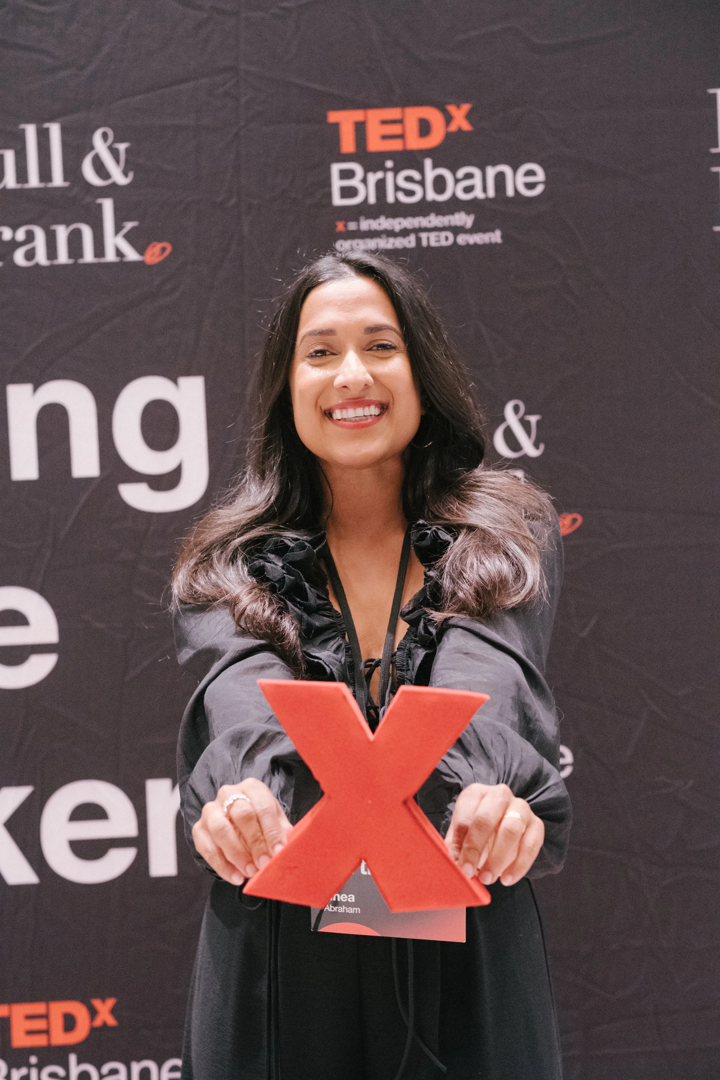 A woman with long dark hair smiling at a TEDx Brisbane event, holding a large red 'X' sign in front of her.