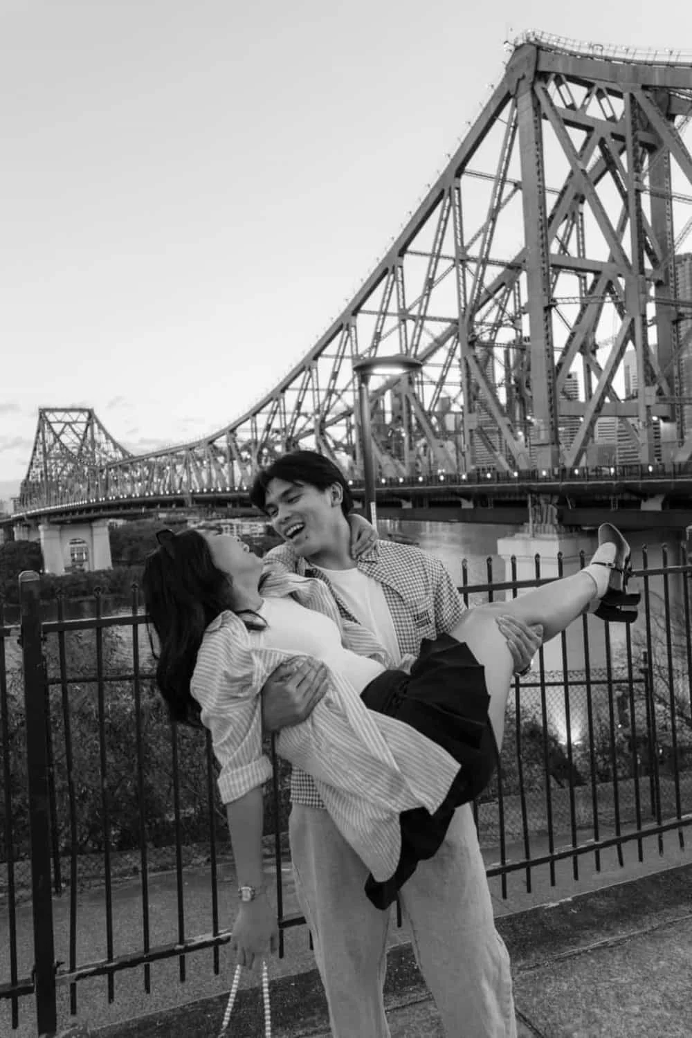 A young man carrying a young woman in his arms, smiling at each other, with a bridge in the background, in black and white.