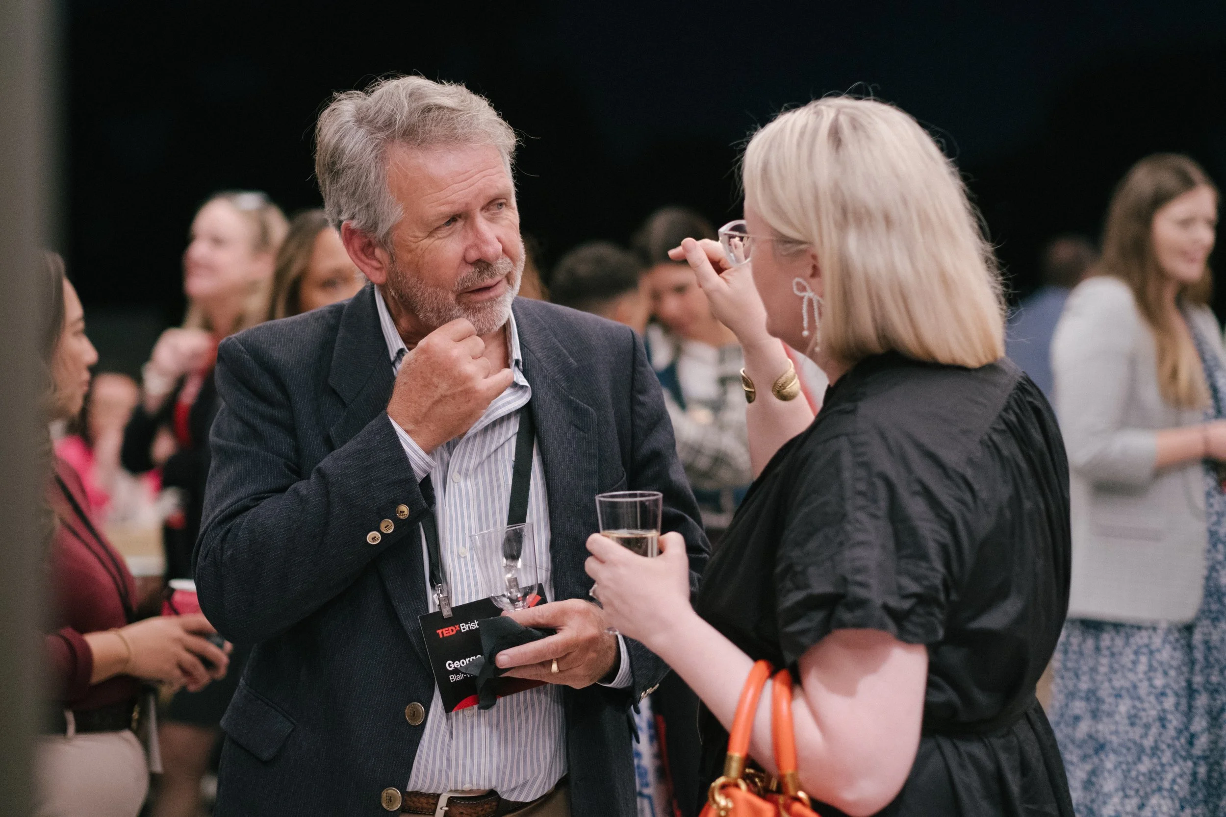 A man with gray hair and a beard wearing a dark blazer converses with a woman with blond hair and glasses at a crowd gathering, both holding glasses of drink.