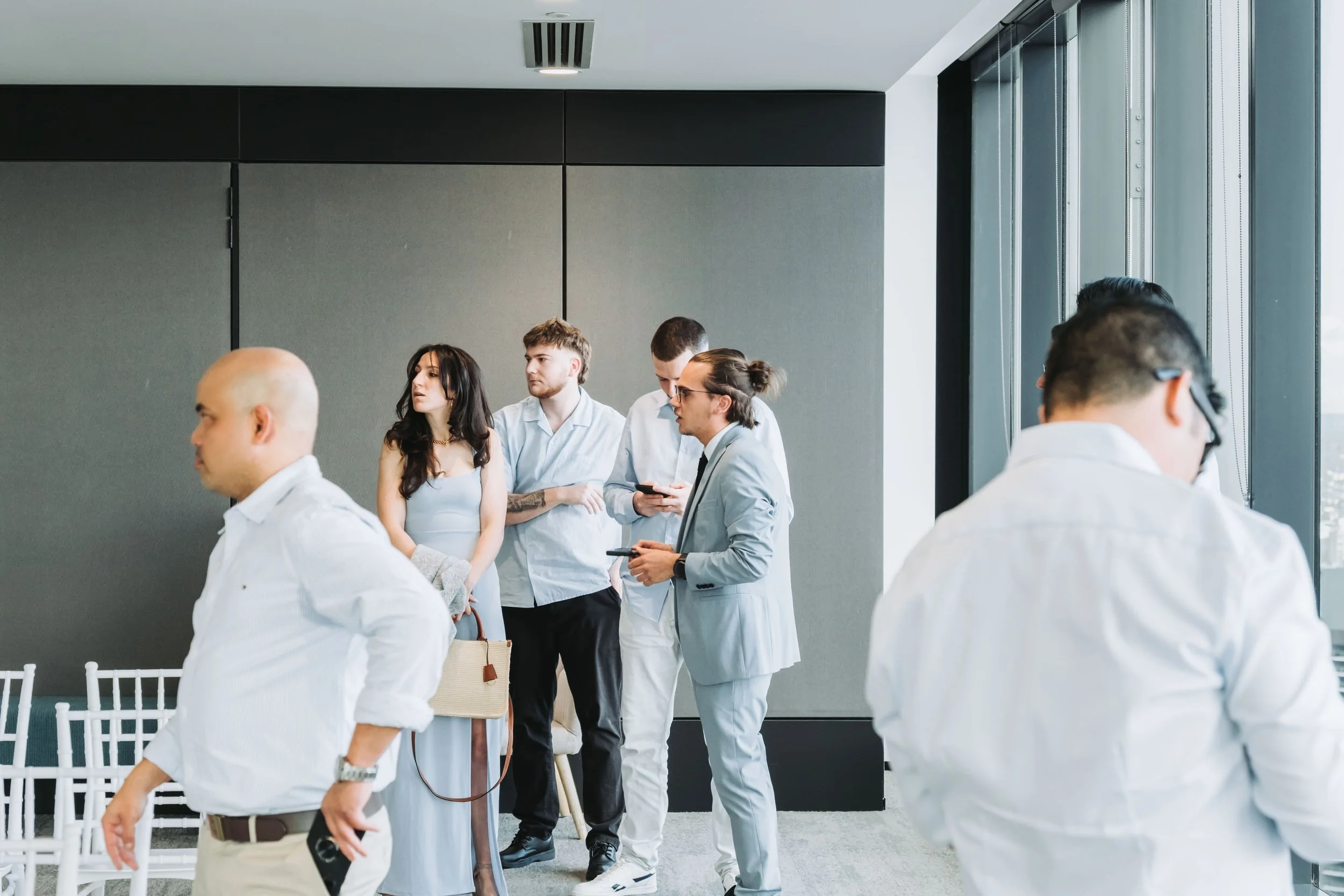 Group of young people dressed in business casual attire standing near a large window in a modern office or conference room.
