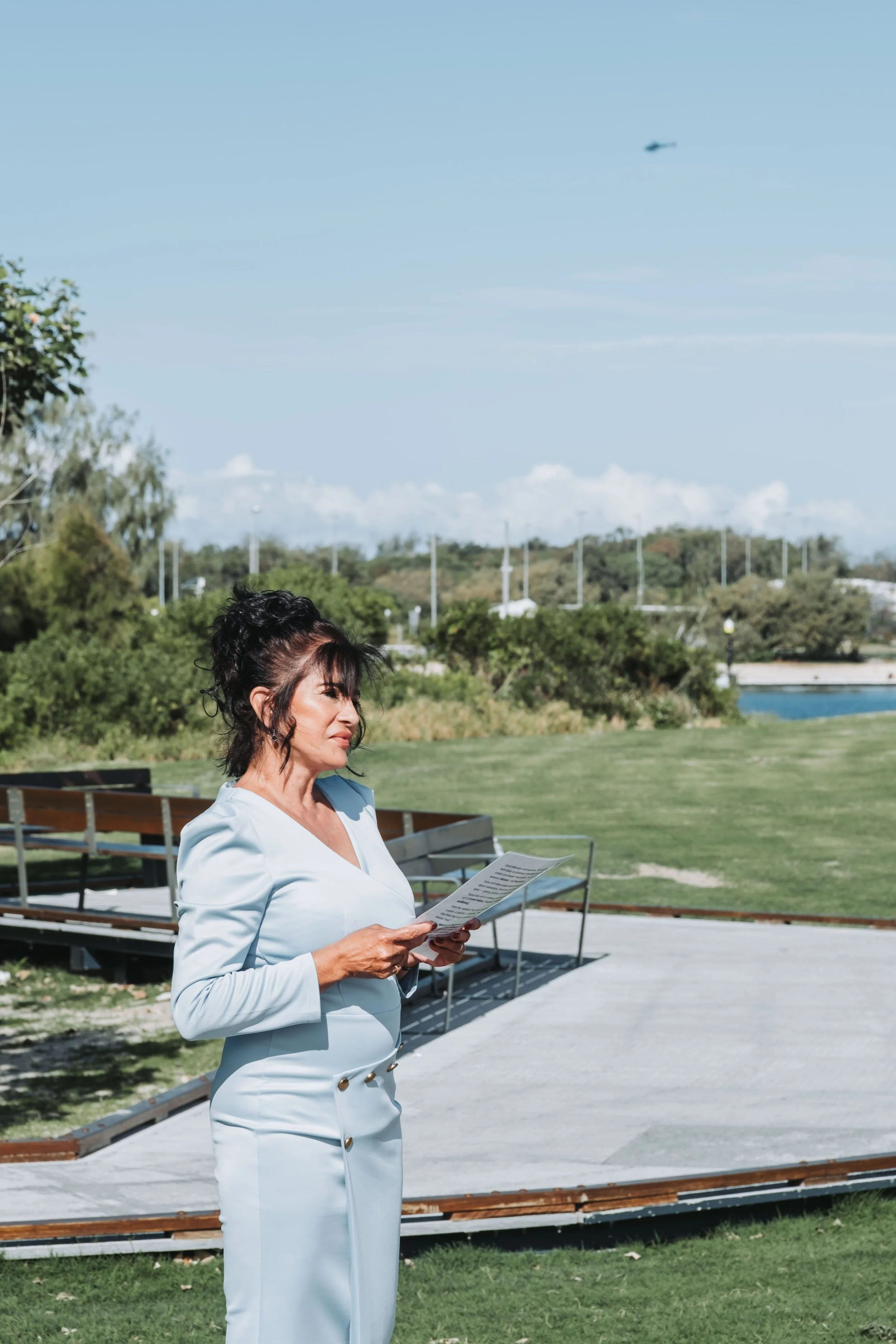 A woman in a white blazer and pants holding a paper, standing outdoors near benches, with greenery and a river in the background, and a small helicopter flying in the sky.