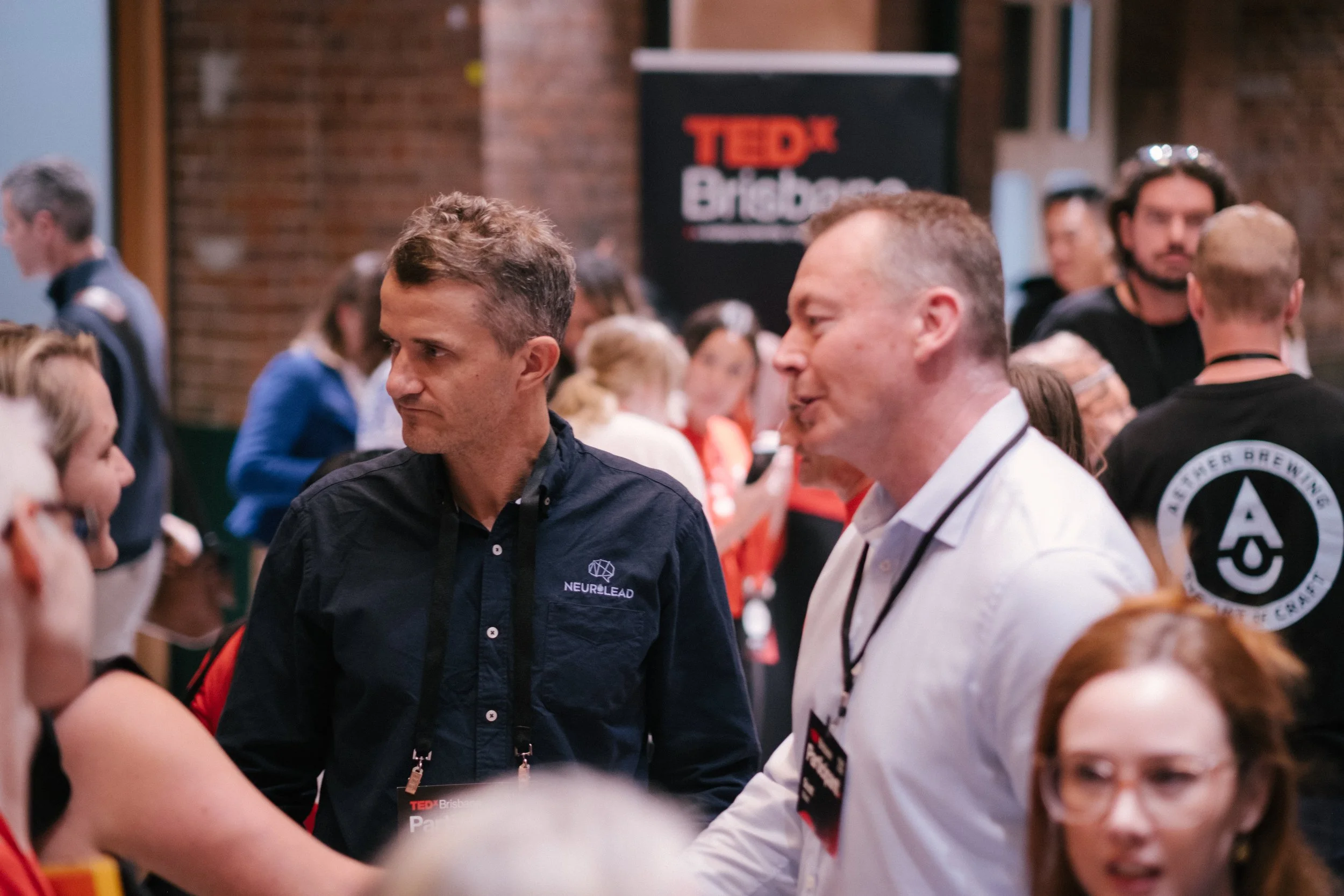 People gathered at a TEDx event, engaging in conversation and networking indoors in front of a black TEDx sign.