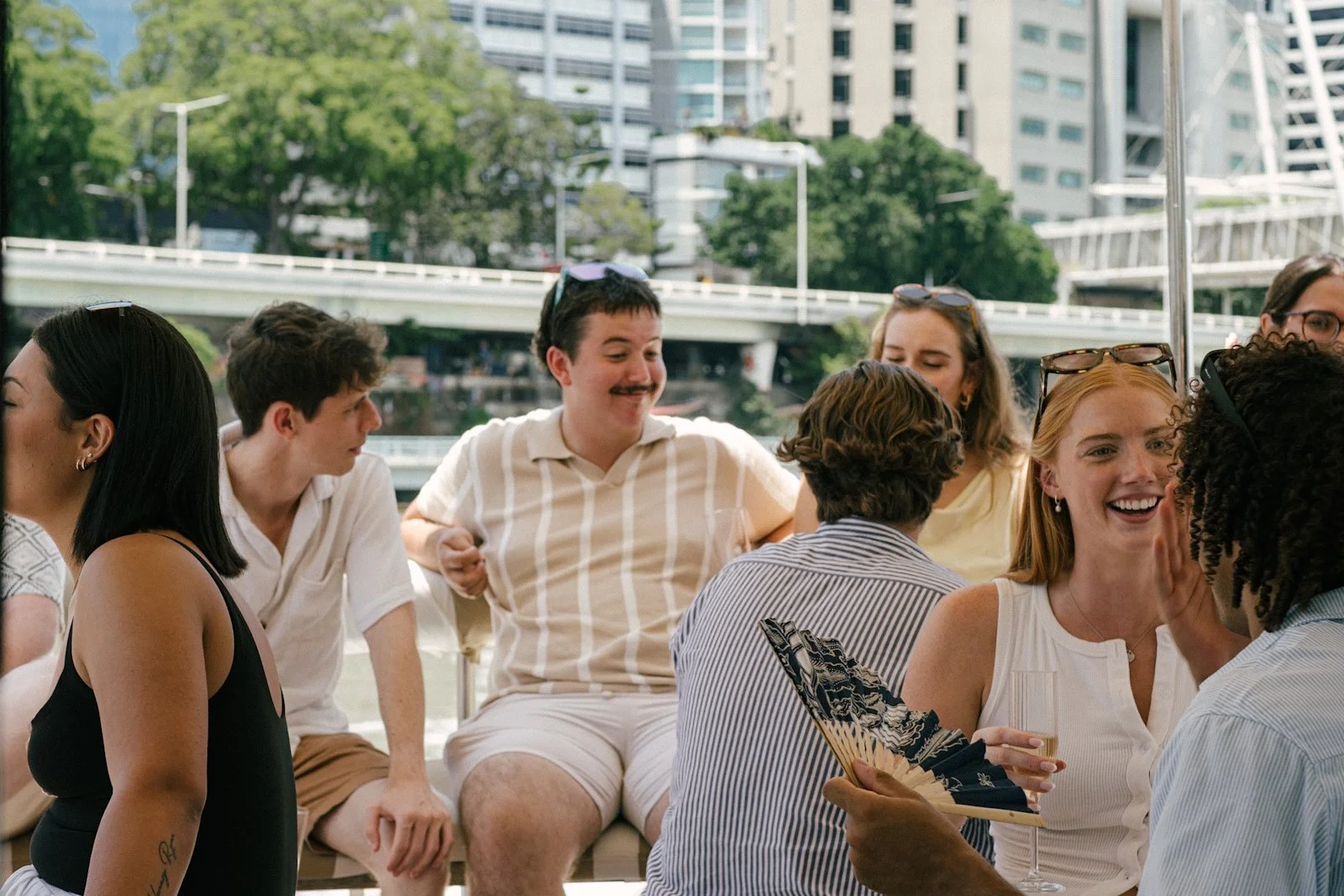 A group of diverse young adults socializing outdoors at a daytime gathering, with a cityscape and bridge in the background. They are smiling and engaging in conversation.