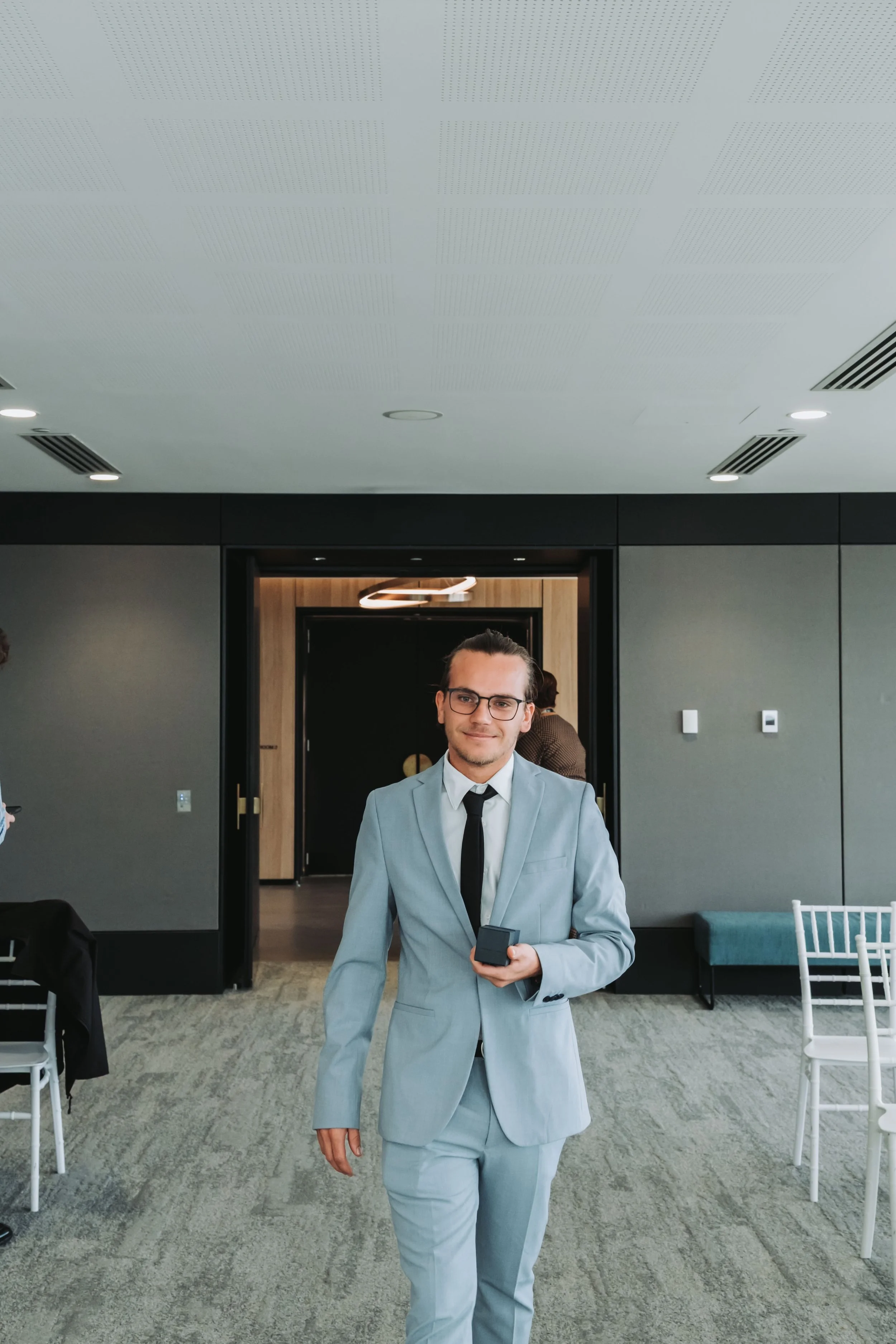 A man dressed in a light gray suit, white shirt, and black tie, holding a small black box, standing inside a modern building with gray walls, white chairs, and a gray carpet. The man is smiling at the camera.