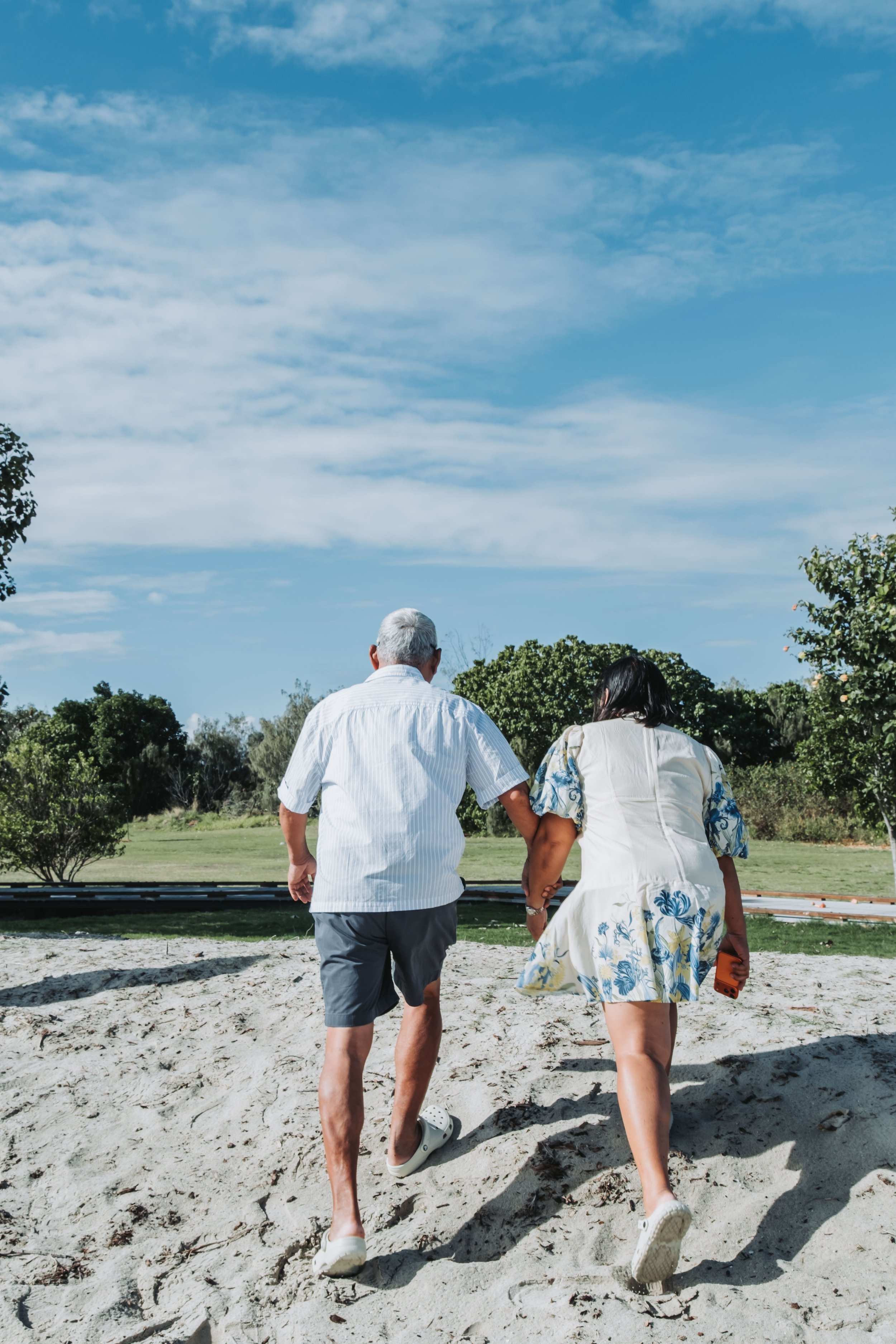Two people walking hand in hand on a sandy area outdoors with trees and a blue sky with clouds in the background.
