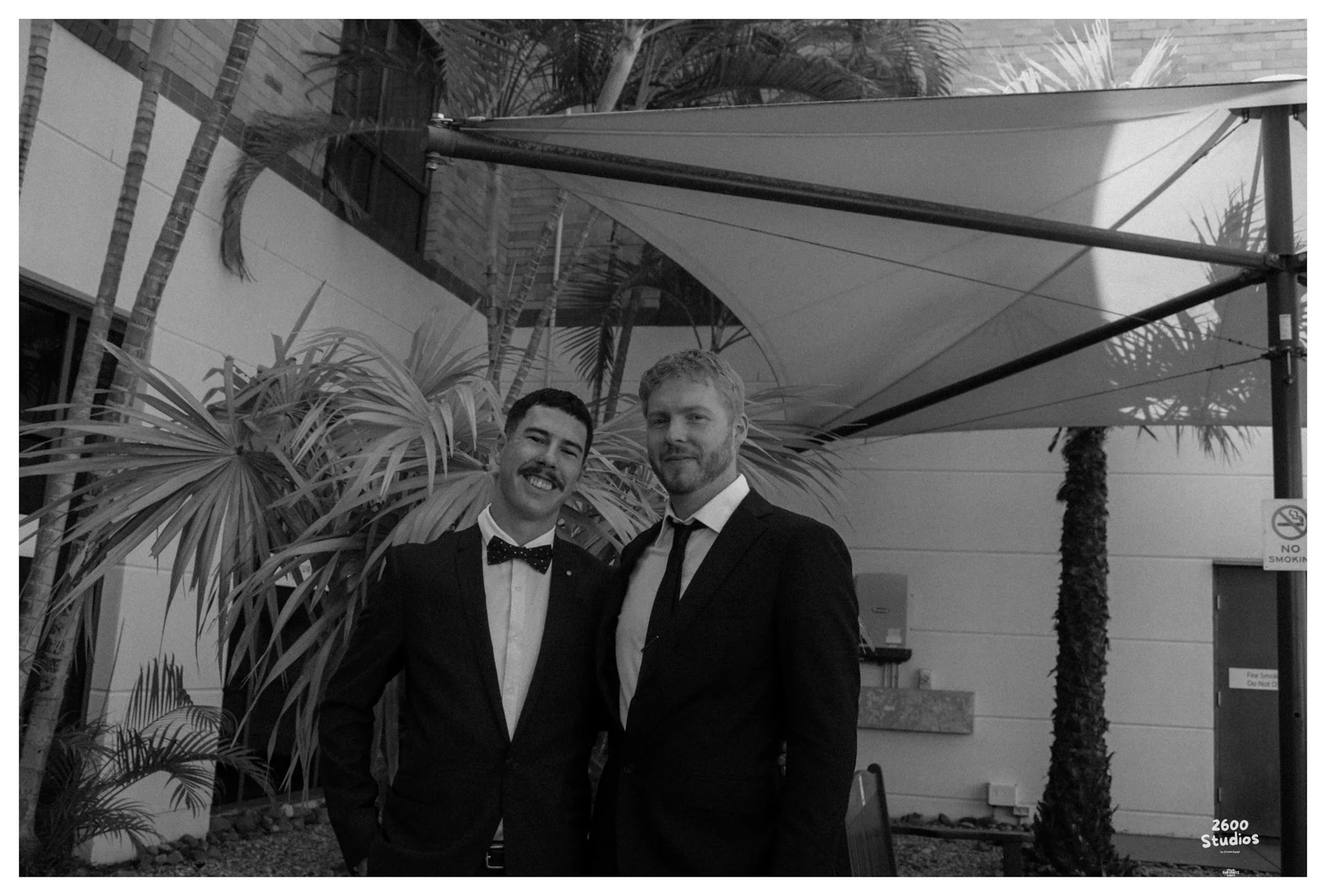 Two men in formal suits and bow ties standing together outdoors, smiling at the camera. Behind them, there are palm trees and a large umbrella, with a building in the background.