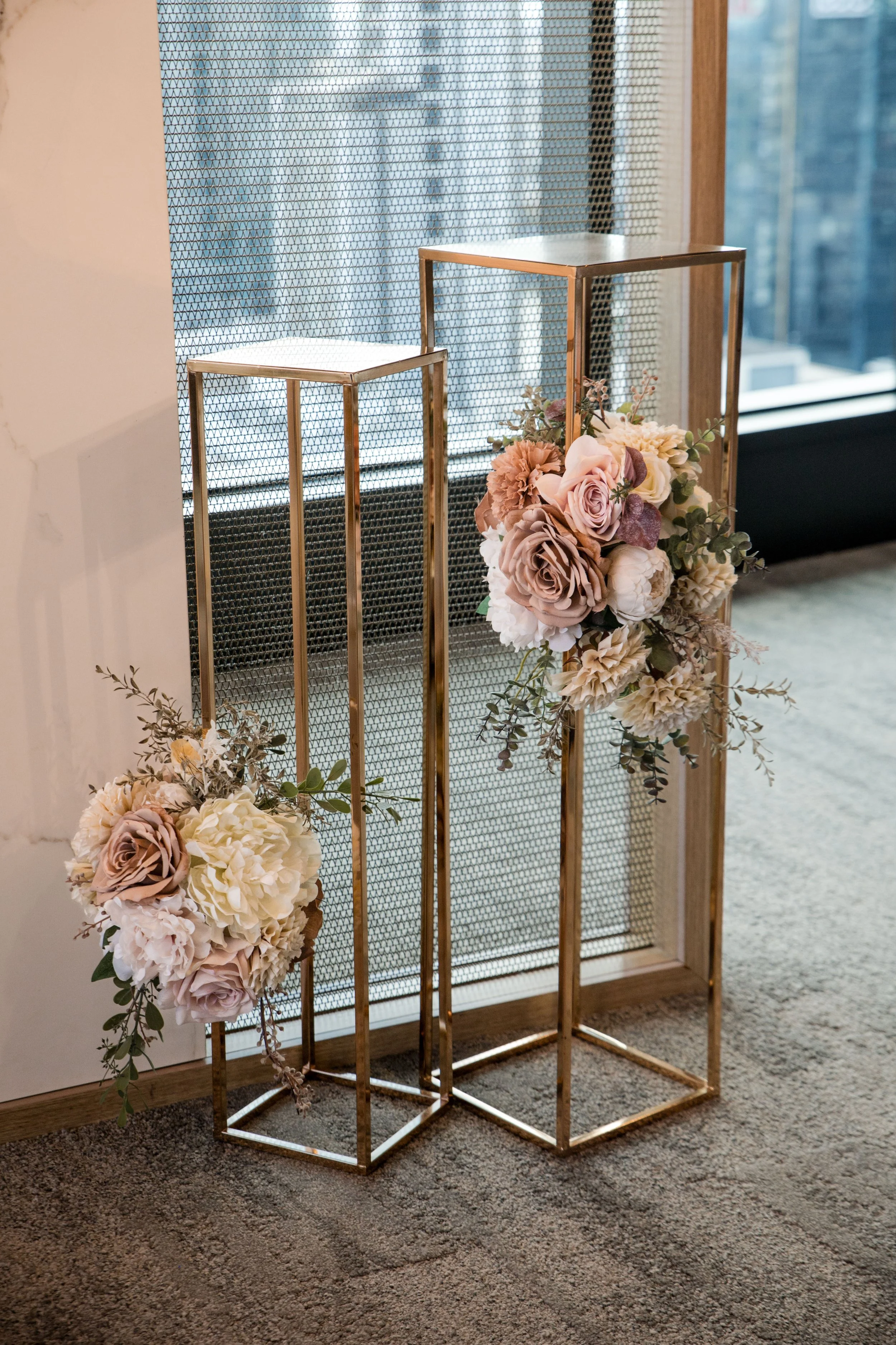 Two gold-colored metal frames with floral arrangements at the base, placed on a carpeted floor near a window with a city view, likely for wedding or event decor.