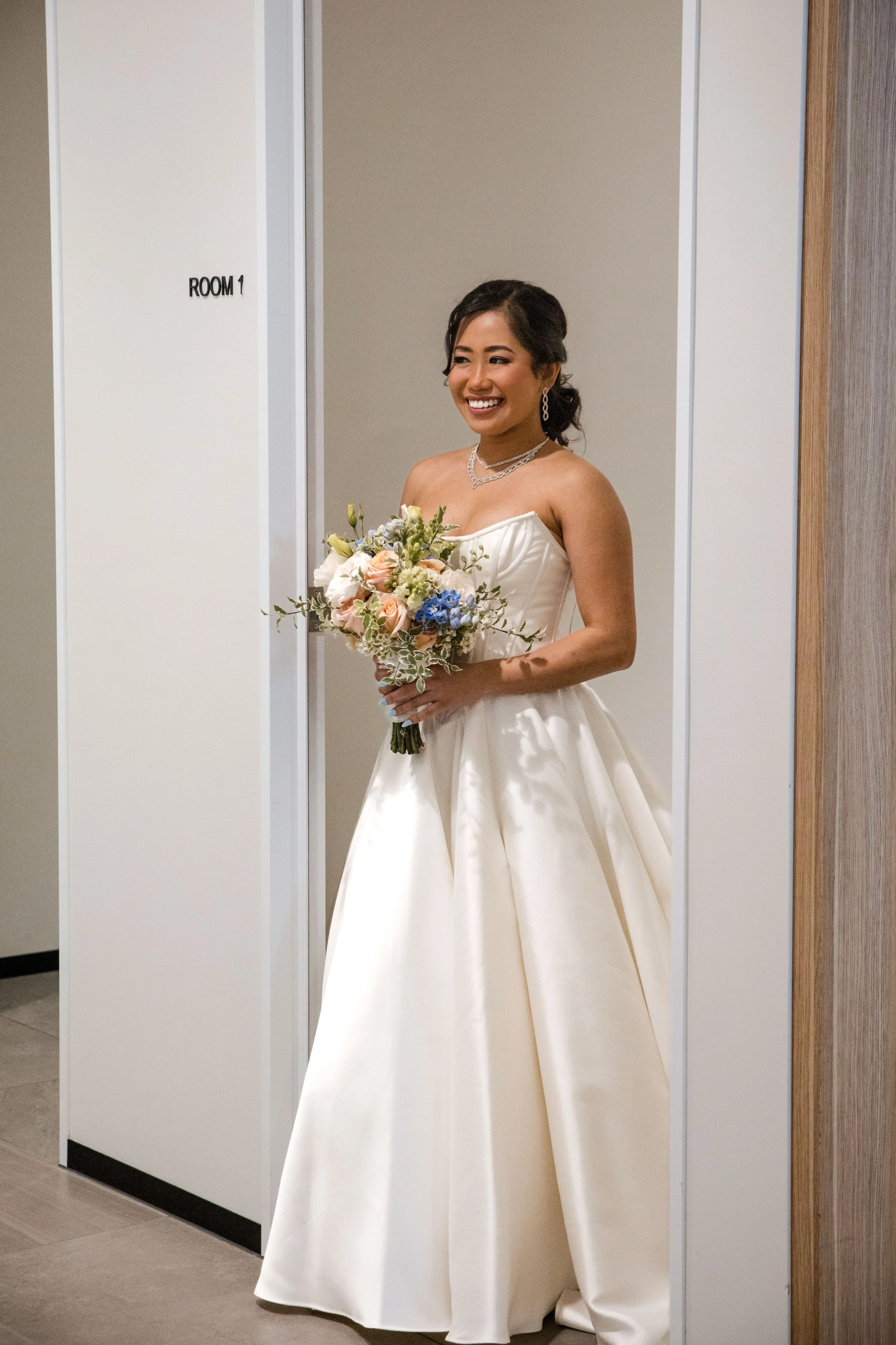 A woman in a white wedding dress holding a bouquet of flowers, smiling, standing in front of a door labeled 'Room 1' in a hallway.