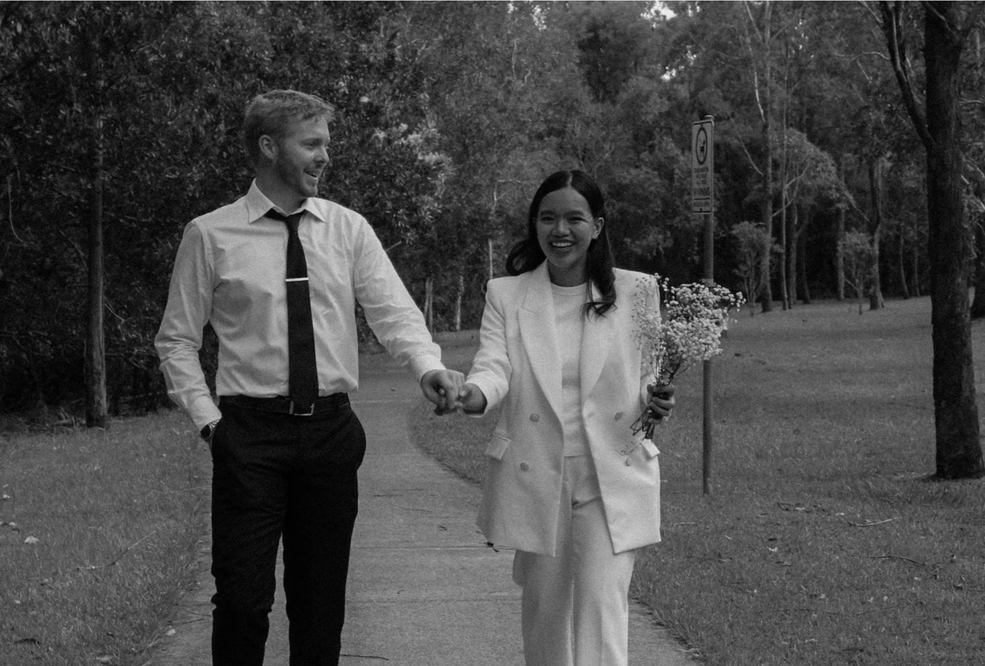 Black and white photo of a couple holding hands on a tree-lined path, woman in white suit holding flowers.