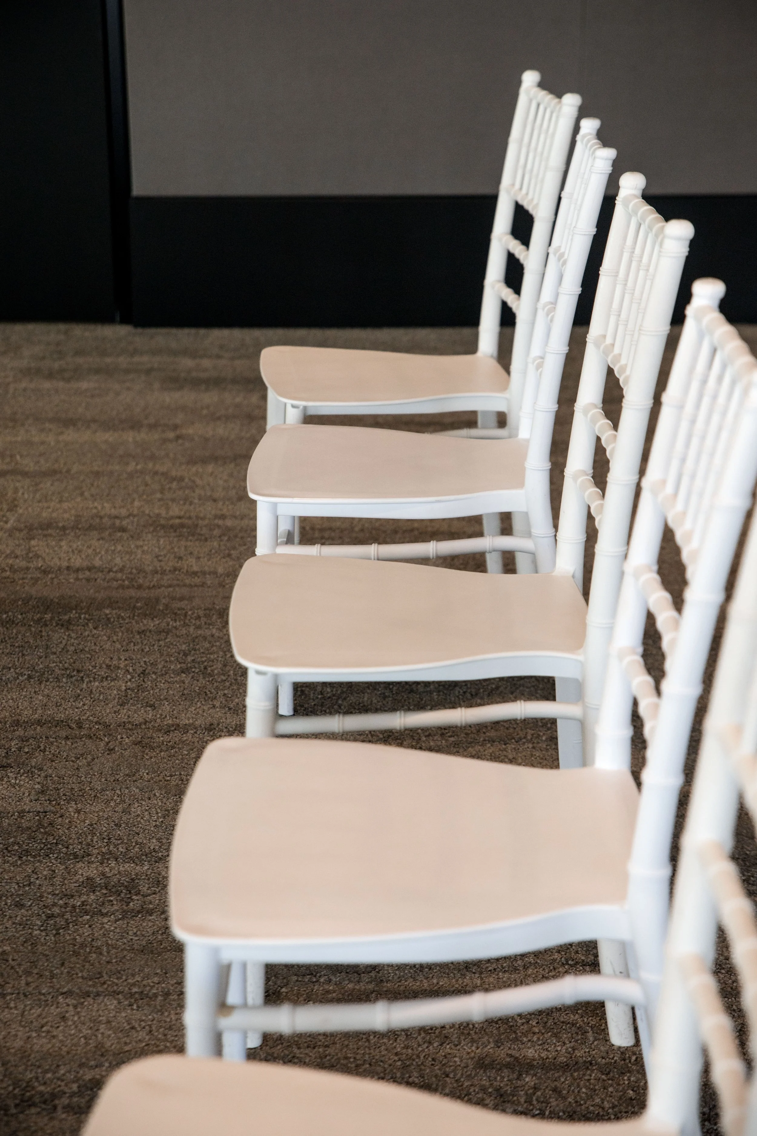 A row of white chairs with beige seats arranged on a brown carpeted floor, with a black and gray wall behind.