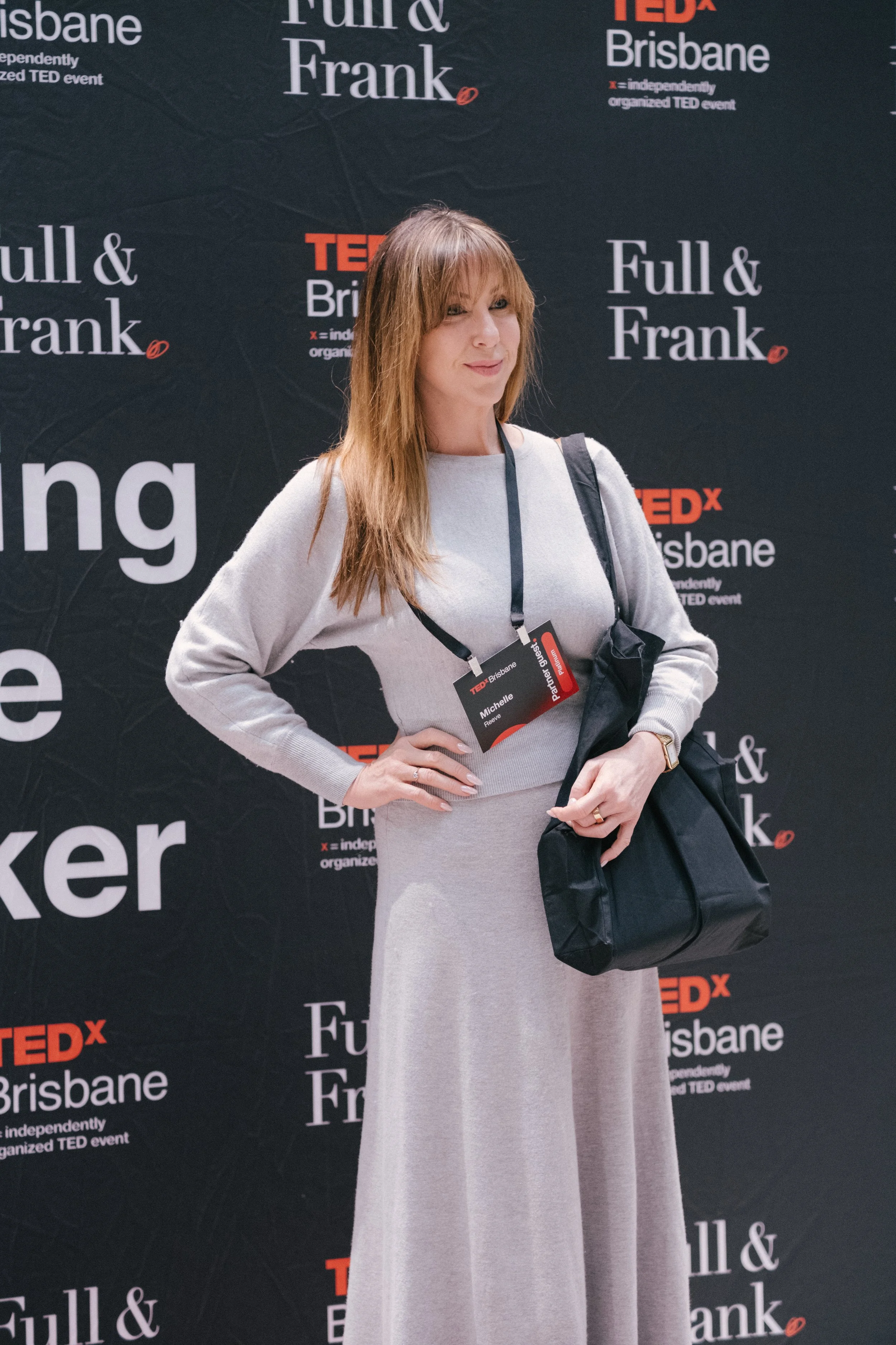 A woman with long reddish-brown hair wearing a light grey dress and a name tag, standing in front of a black backdrop with the TEDx Brisbane logo and the words "Full & Frank".