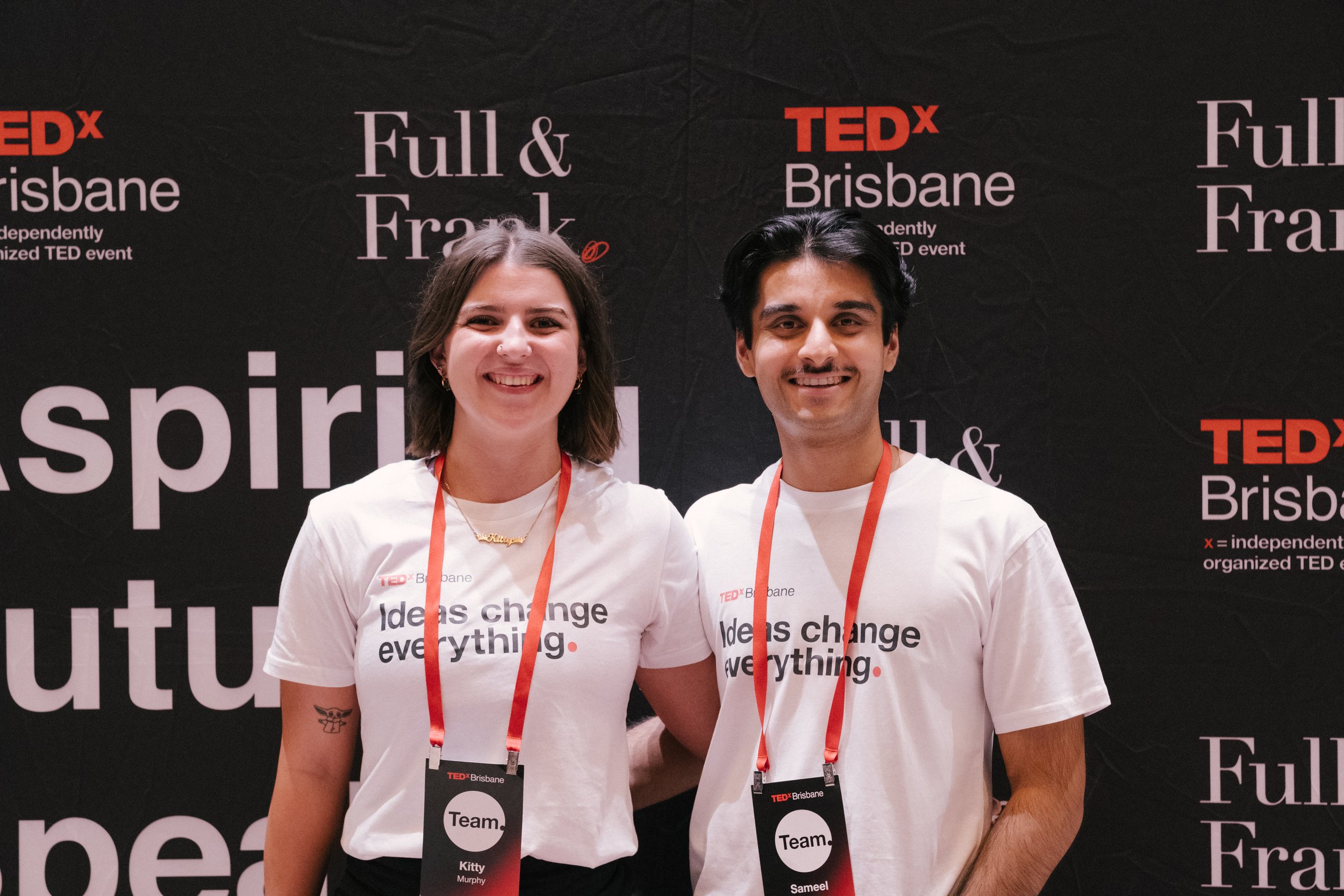 Two people standing in front of a black TEDx Brisbane backdrop, wearing white t-shirts with the phrase 'ideas change everything.' Each has a microphone badge around their neck, indicating they are part of a team at the event.