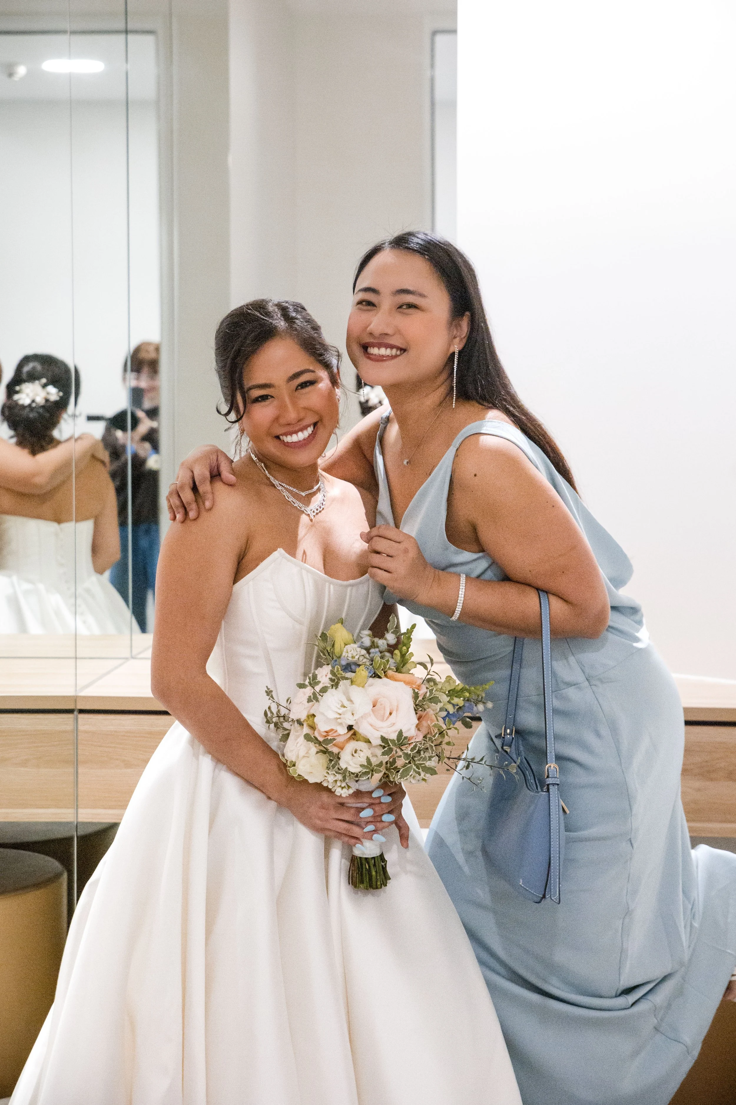 A bride in a white wedding dress and a woman in a light blue dress smiling and hugging at a wedding reception, holding a bouquet of flowers, with a mirror reflecting other guests in the background.