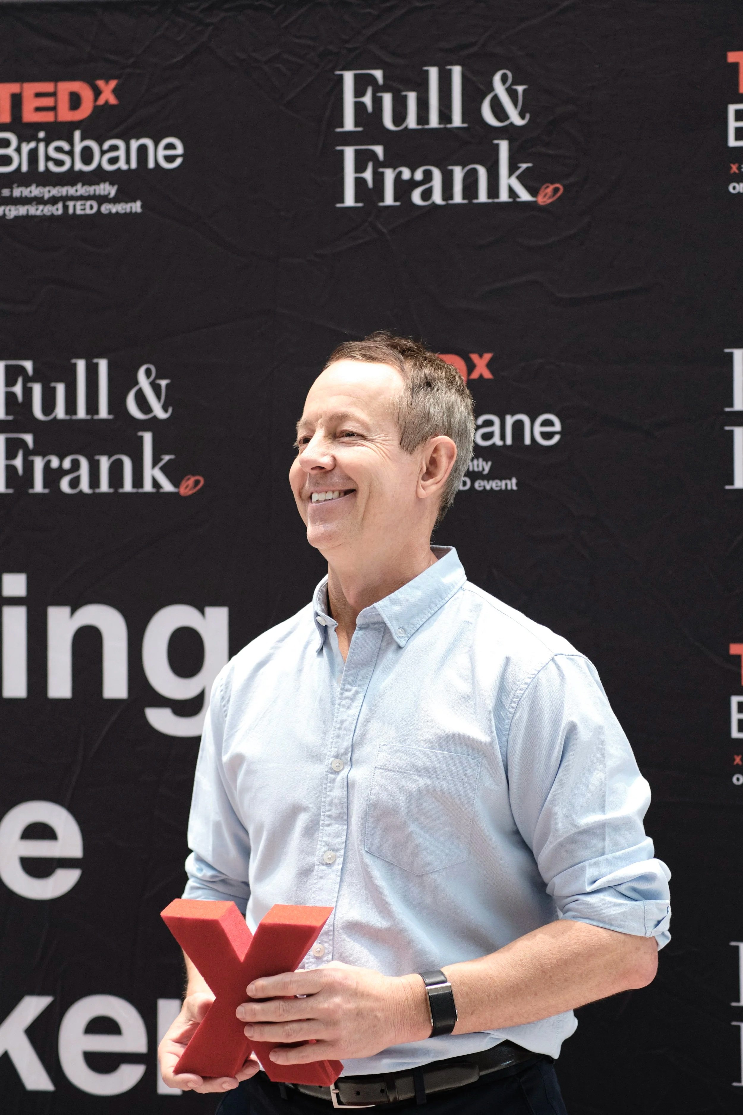 A smiling man in a light blue dress shirt holding a red TEDx 'X' symbol in front of a black backdrop with TEDx Brisbane and Full & Frank written on it.