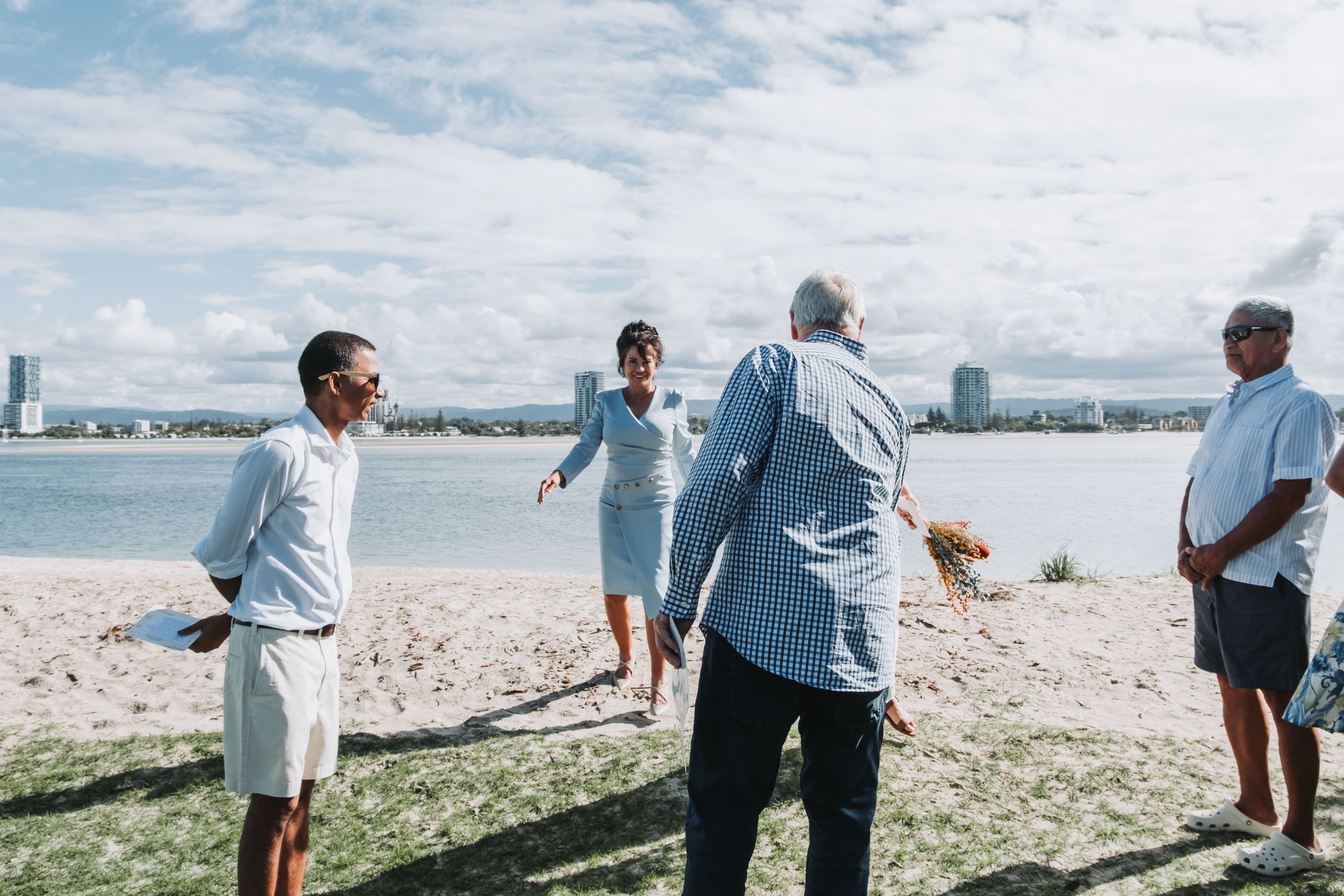A group of five people standing on a sandy beach, engaging in conversation during daytime with a city skyline and water in the background.