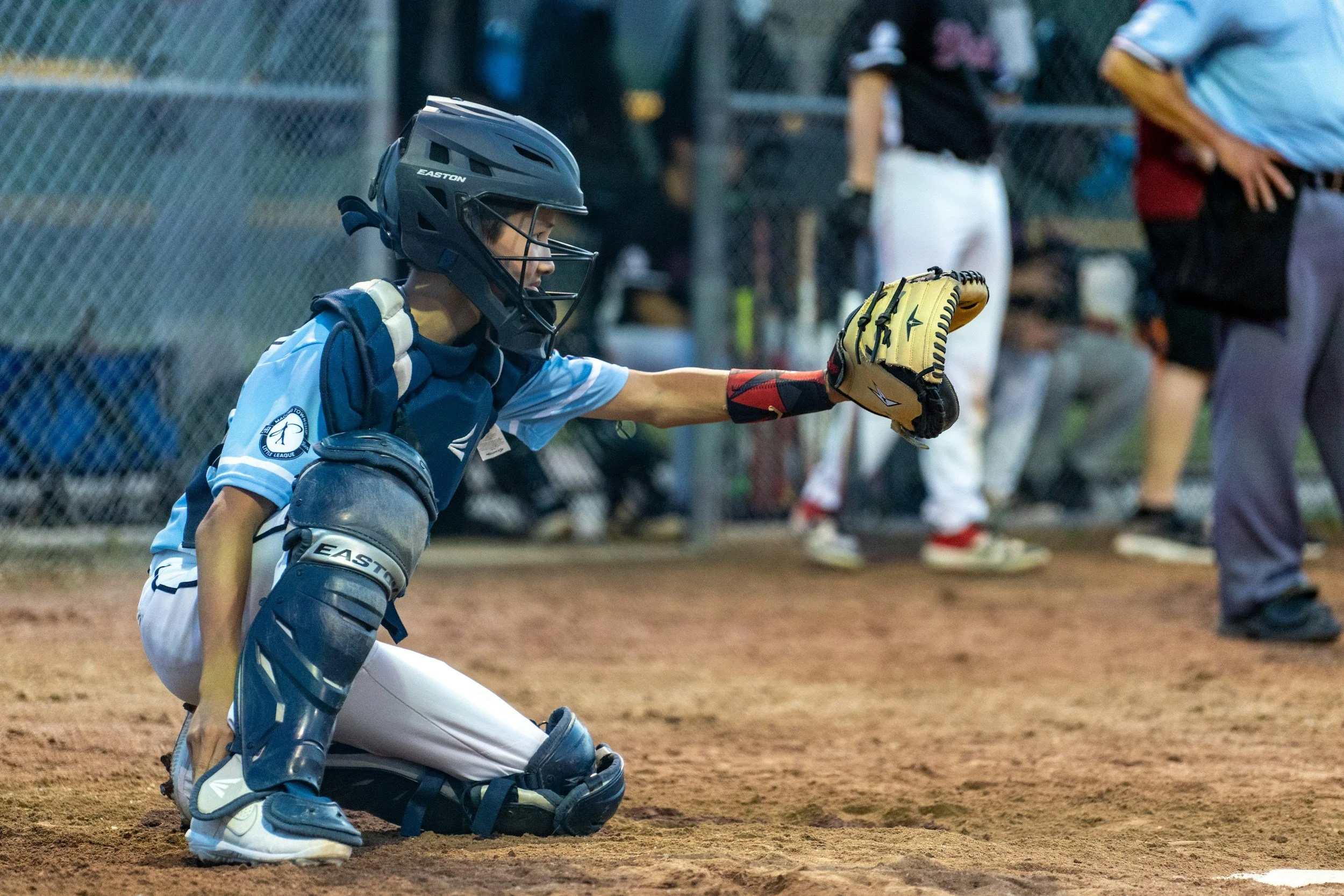 A young baseball catcher in protective gear, kneeling on the dirt field with a baseball glove extended forward during a game.