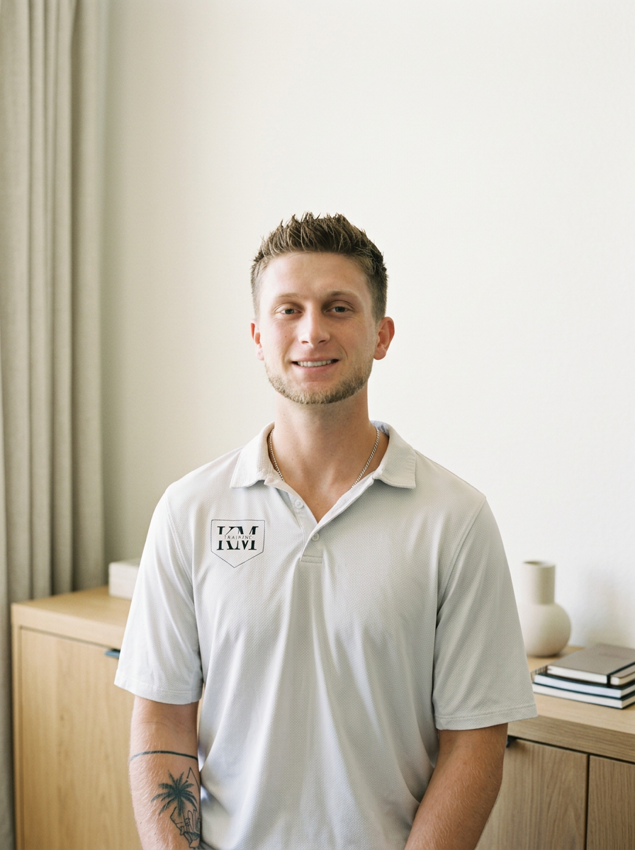 A young man with short, styled hair and a beard, smiling while standing in a room with light-colored walls. He is wearing a white polo shirt with a logo on the left side that reads 'TAM' and 'Training'. The room has a wooden dresser with a few books and a decorative white vase on top, and curtains on the left side.