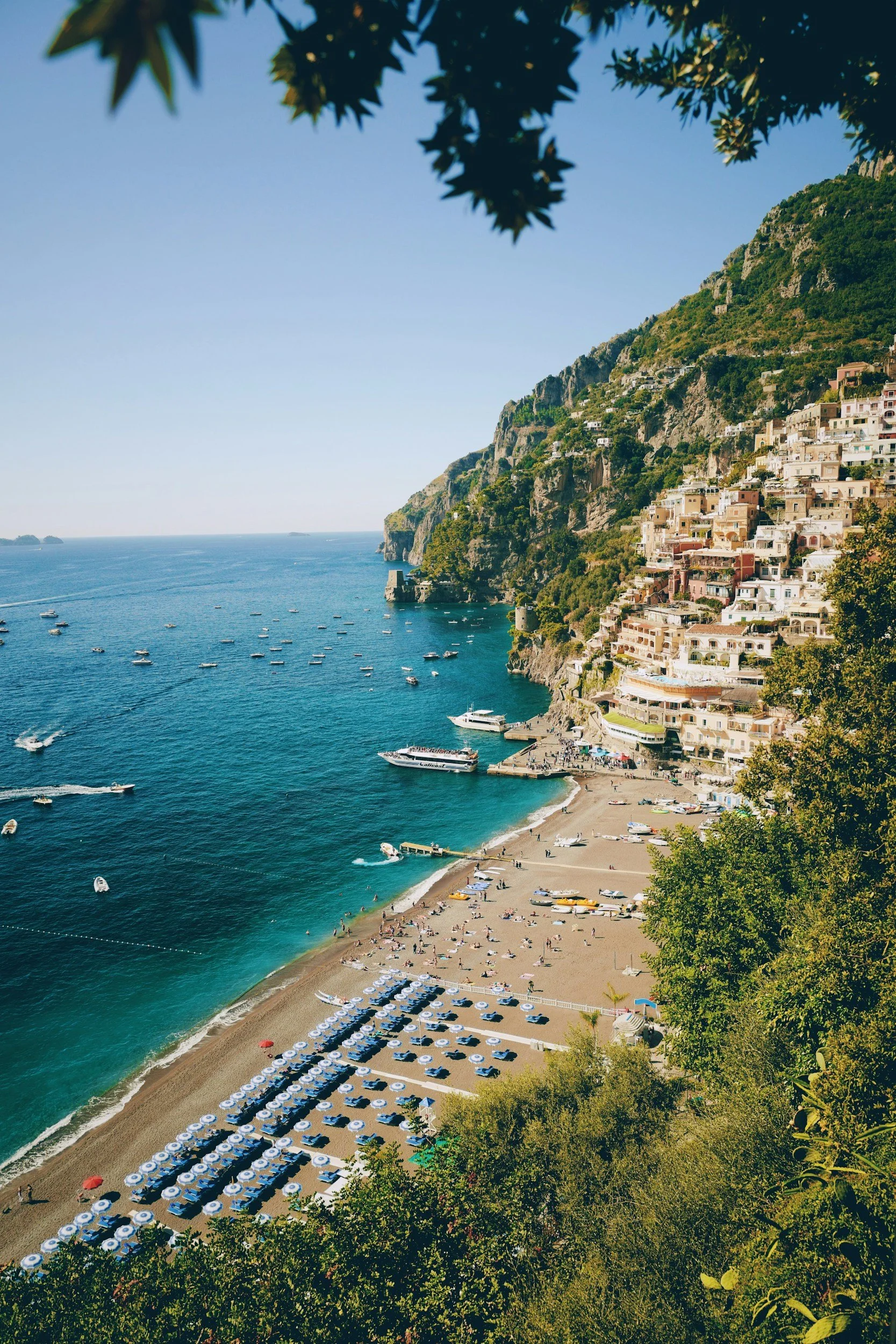 A picturesque coastal scene of a beach with sun umbrellas and lounge chairs, turquoise water with boats, and a hillside town with colorful buildings nestled into green cliffs, under a clear blue sky.