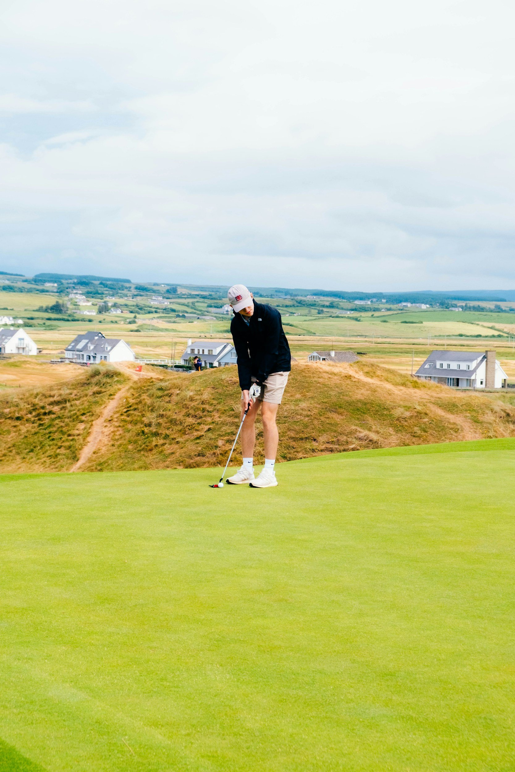 A person preparing to putt on a golf green with a scenic landscape and houses in the background.