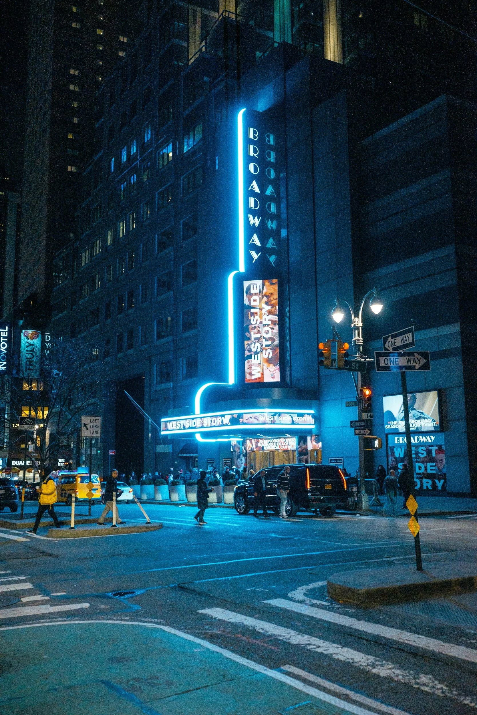 Night scene of Times Square in New York City with a lit Broadway sign, digital billboards, pedestrians crossing the street, taxis, and cars, with high-rise buildings in the background.