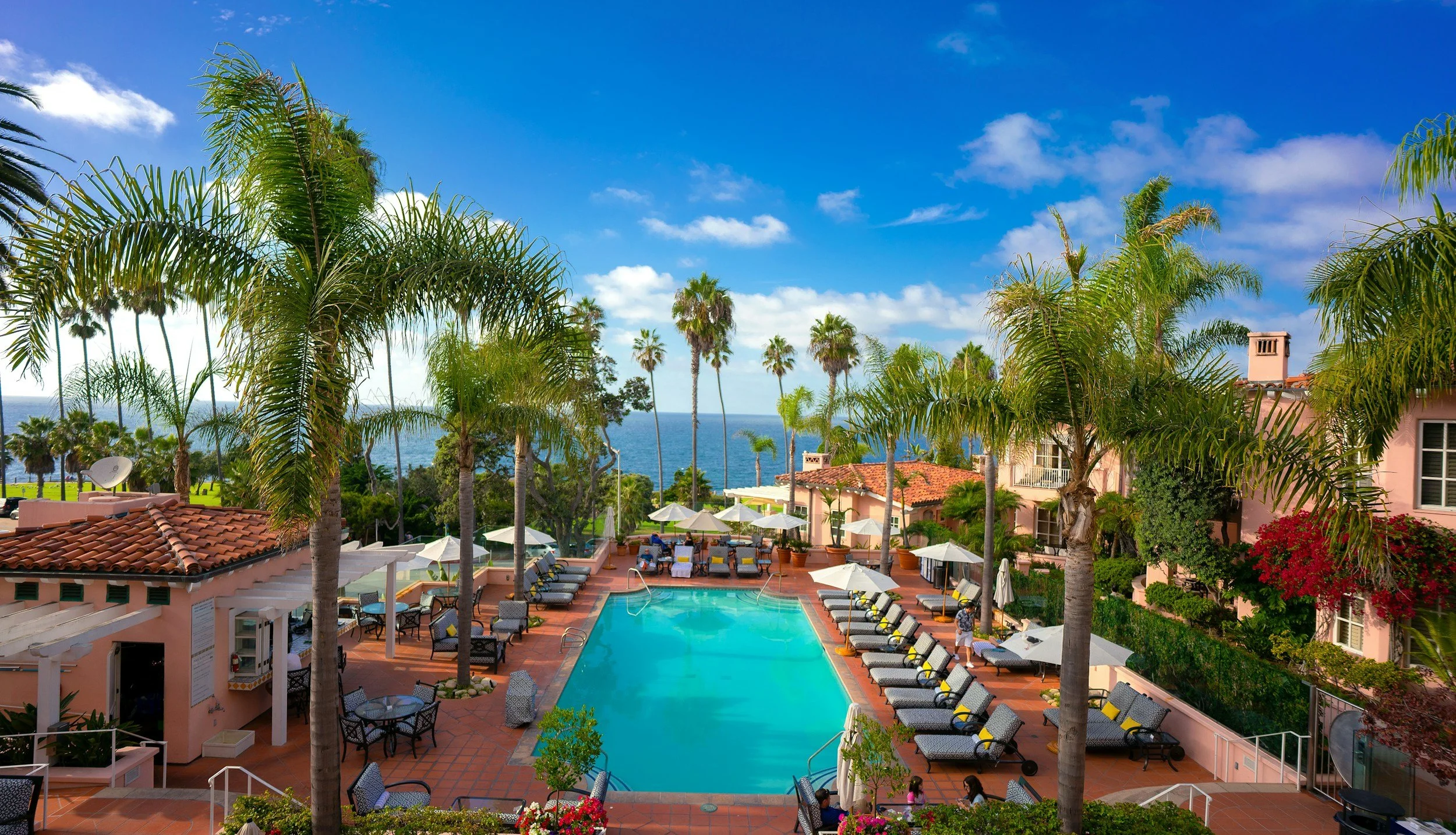 A resort with a swimming pool surrounded by lounge chairs and umbrellas, tall palm trees, and a scenic view of the ocean in the background under a blue sky with some clouds.
