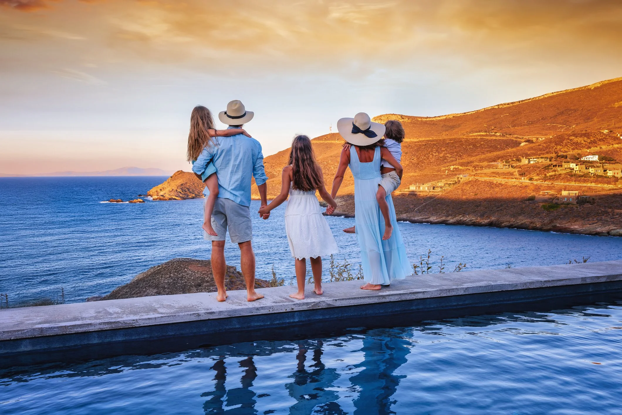 A group of people standing together on the beach, facing the ocean, with some of their arms around each other. The scene shows a cloudy sky and gentle waves on the water.