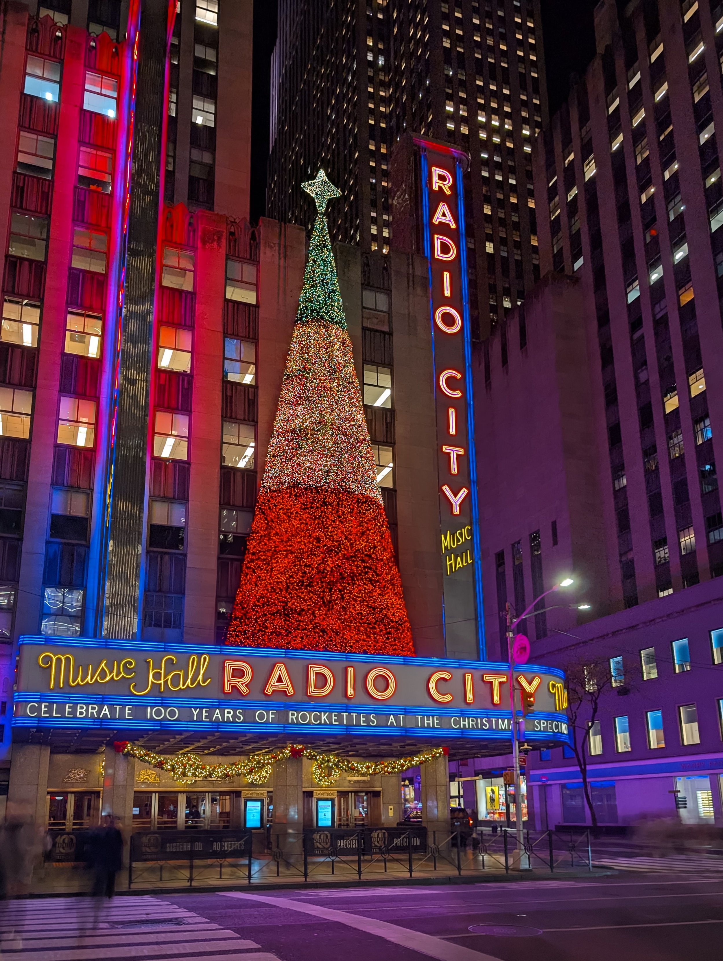 Night scene of Times Square in New York City with a lit Broadway sign, digital billboards, pedestrians crossing the street, taxis, and cars, with high-rise buildings in the background.