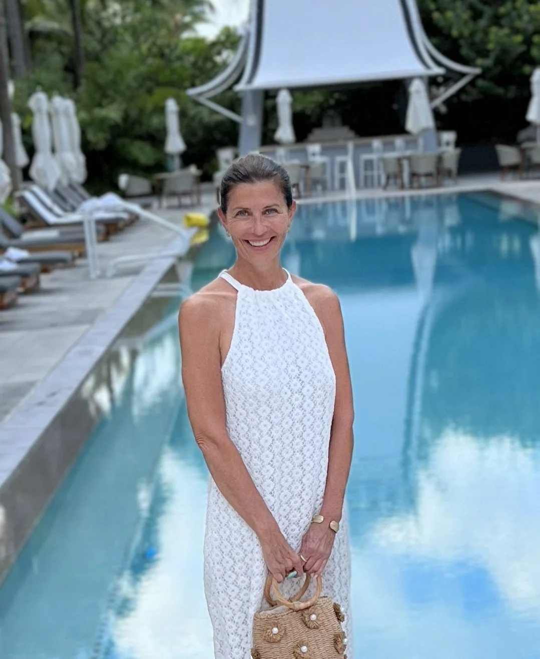 Smiling woman in a white lace dress standing poolside at a resort or hotel, holding a straw handbag, with lounge chairs and umbrellas in the background.