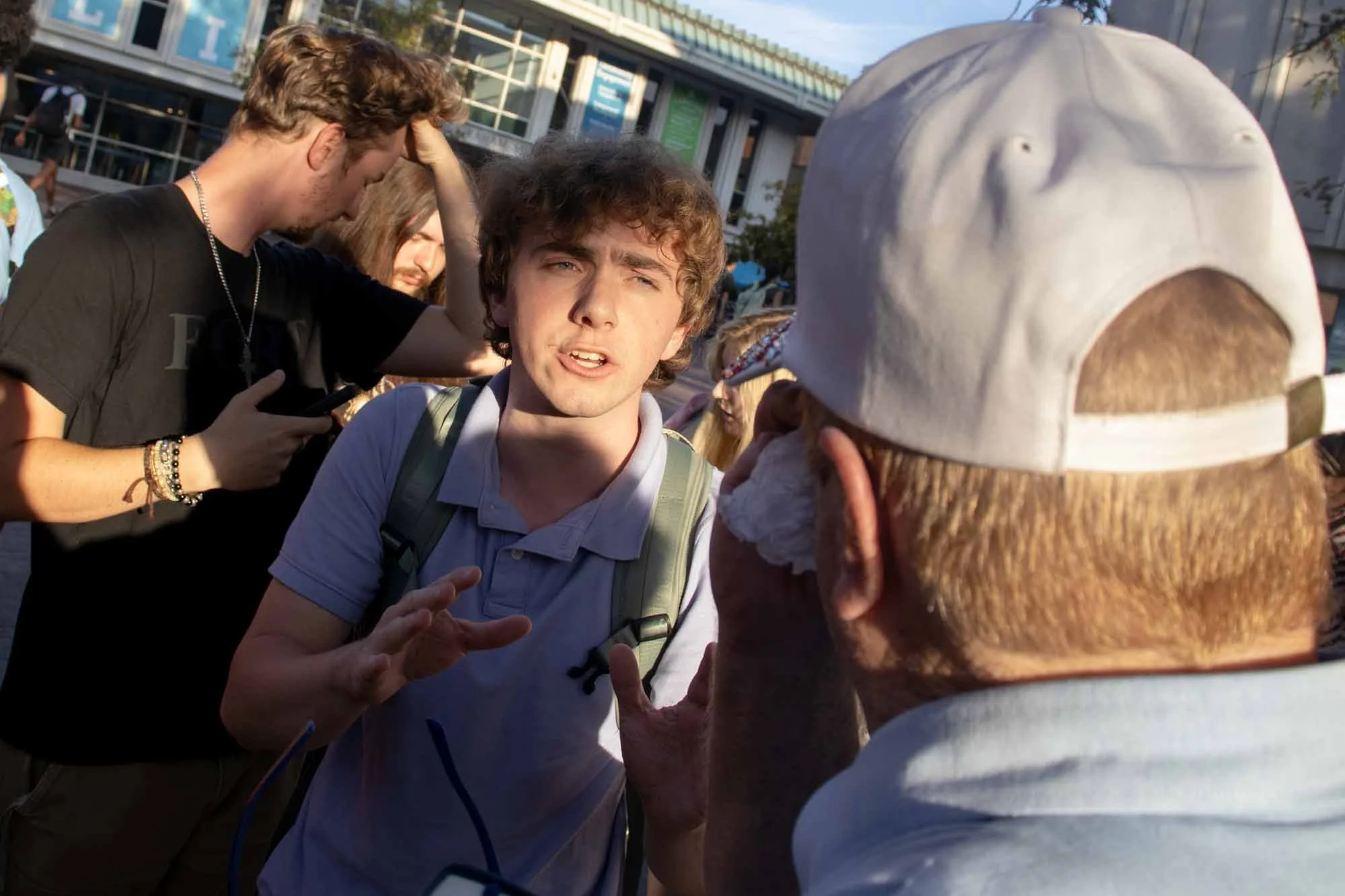 A young man with brown curly hair, wearing a white polo shirt with a backpack, appears to be talking to an elderly person with a white baseball cap. There are other people around, including a man in a black shirt with a chain necklace and a girl with