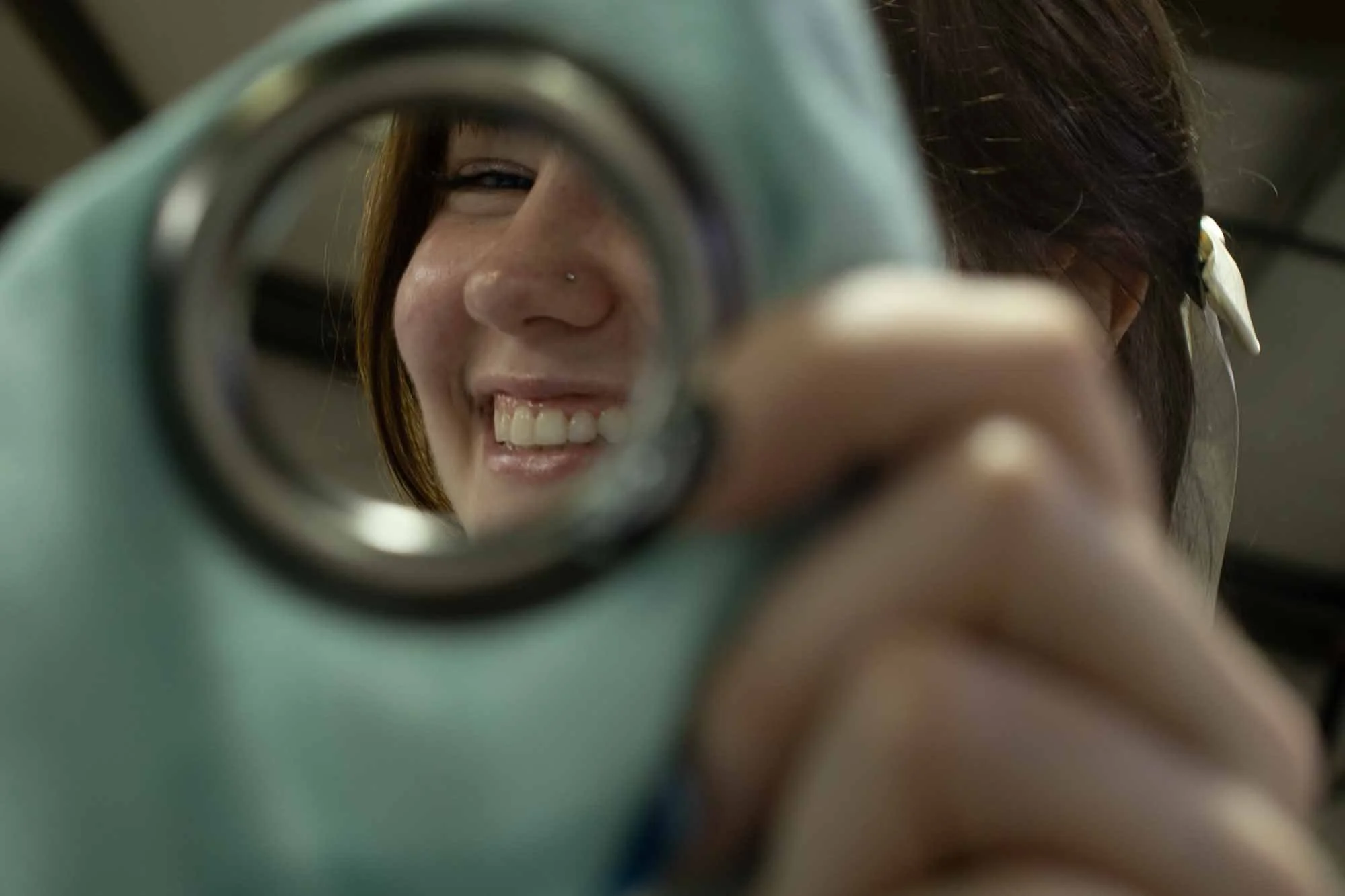 A person with short reddish-brown hair and a nose piercing smiling while looking into a green oscilloscope.