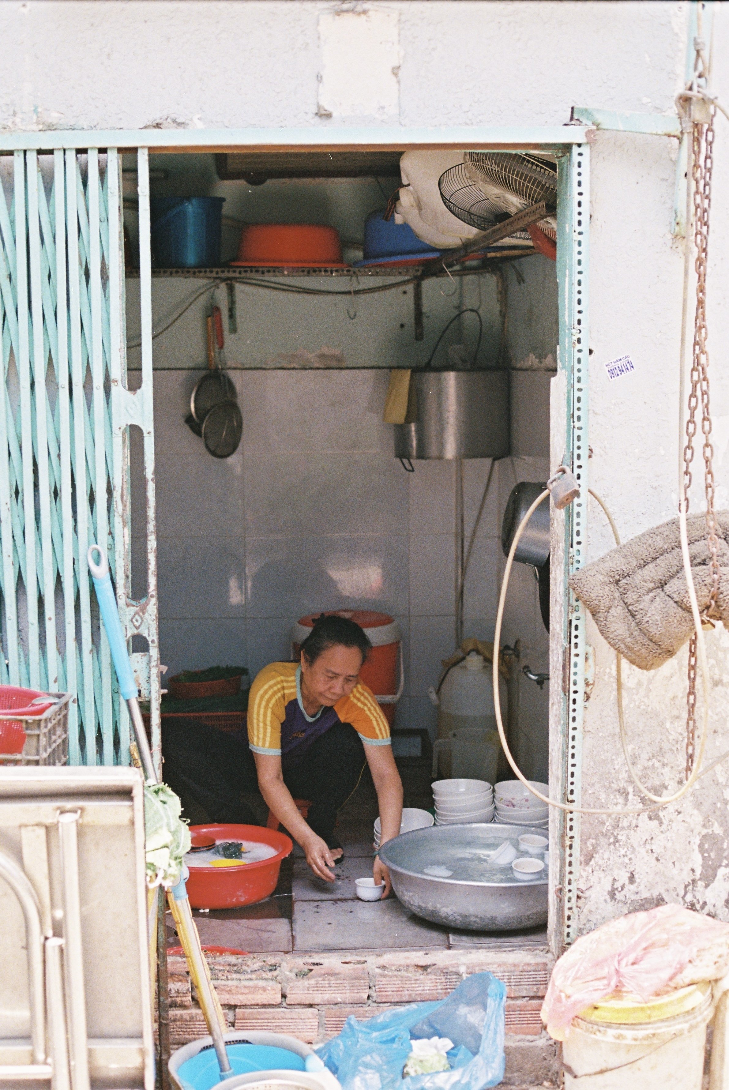 A woman squatting and washing dishes in a small outdoor kitchen, with various kitchen utensils, containers, and water basins around her.