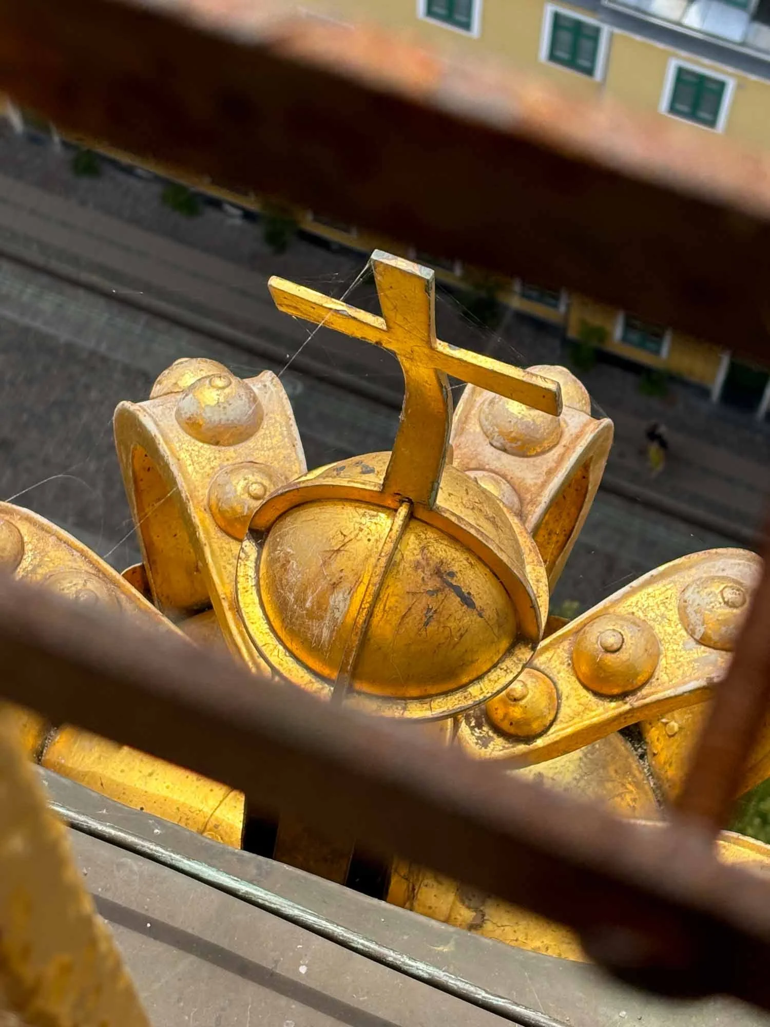 Close-up view of a decorative yellow metal structure, possibly part of a fence or railing, with ornate design and some cobwebs, on an outdoor building with yellow walls and green window frames in the background.