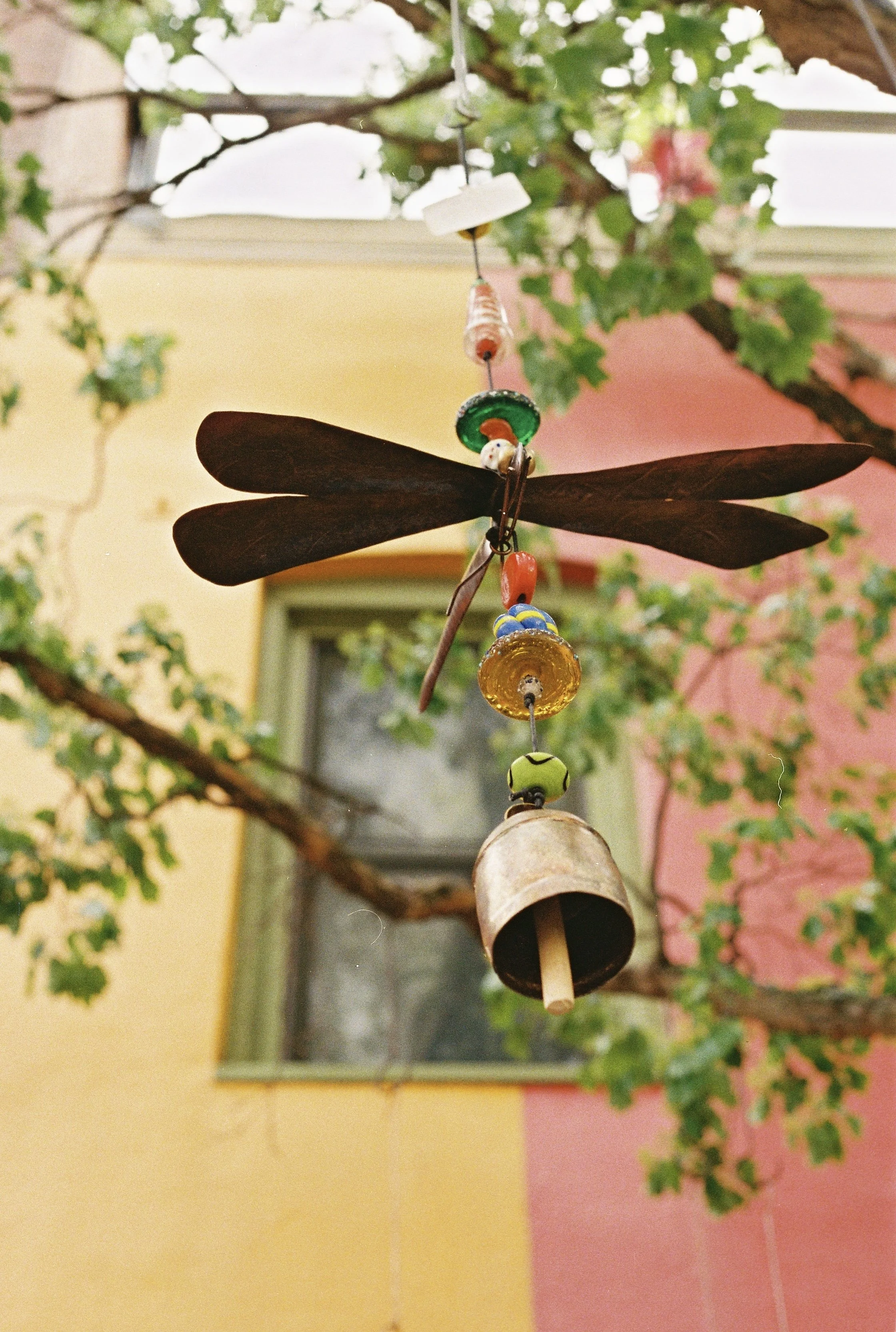 A hanging mobile with a large wooden propeller, colorful glass beads, and a small bell, suspended from a branch with green leaves, against a background of yellow, pink, and beige walls with a window.