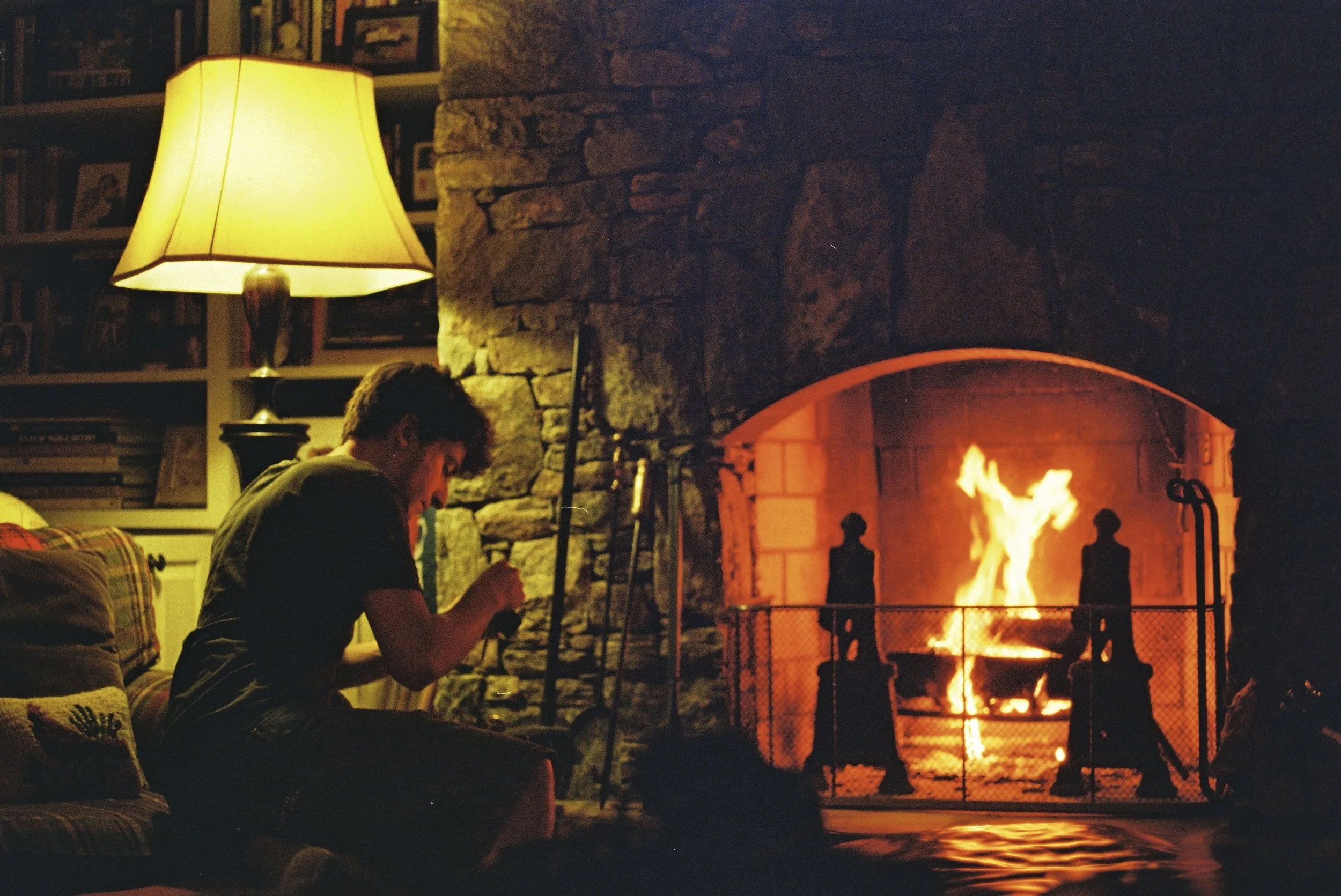 A person sitting on a couch, using their phone, near a fireplace with a bright flame, inside a cozy living room with a yellow lamp and bookshelves.