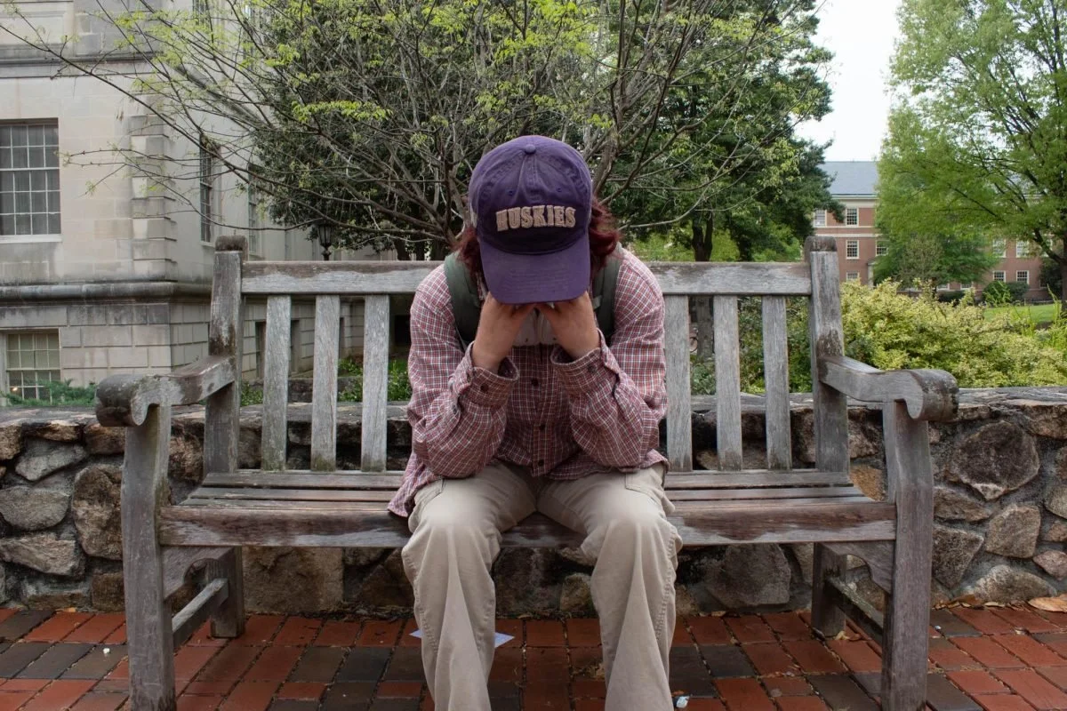 Person sitting on a wooden bench with hands covering face, wearing a purple cap with 'Huskies' embroidered, plaid shirt, and beige pants, outdoors with trees and buildings in the background.