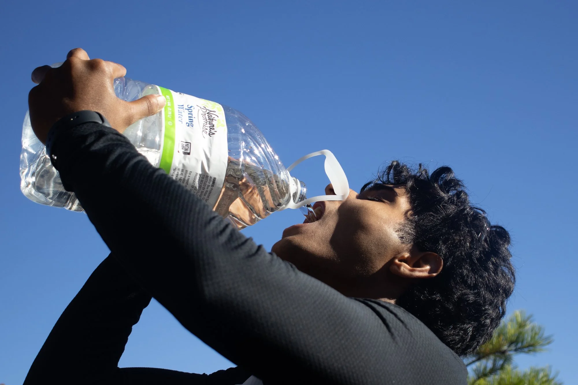 A person with short, dark curly hair and dark skin is holding a large plastic bottle of water and drinking from a white straw outdoors against a bright blue sky.