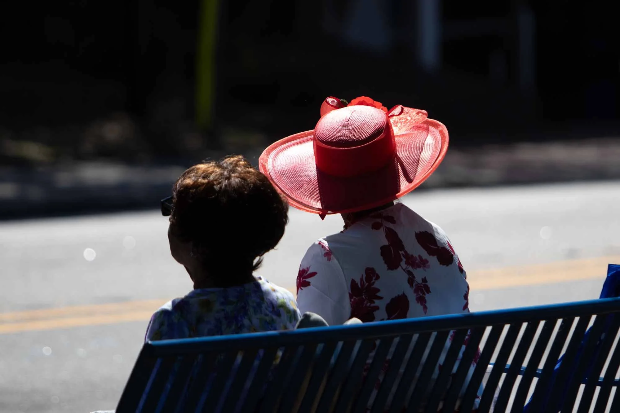 Two women sitting on a bench at an outdoor event, one wearing a large red hat and a floral top, the other with short hair and glasses, with blurred background and sunlight.