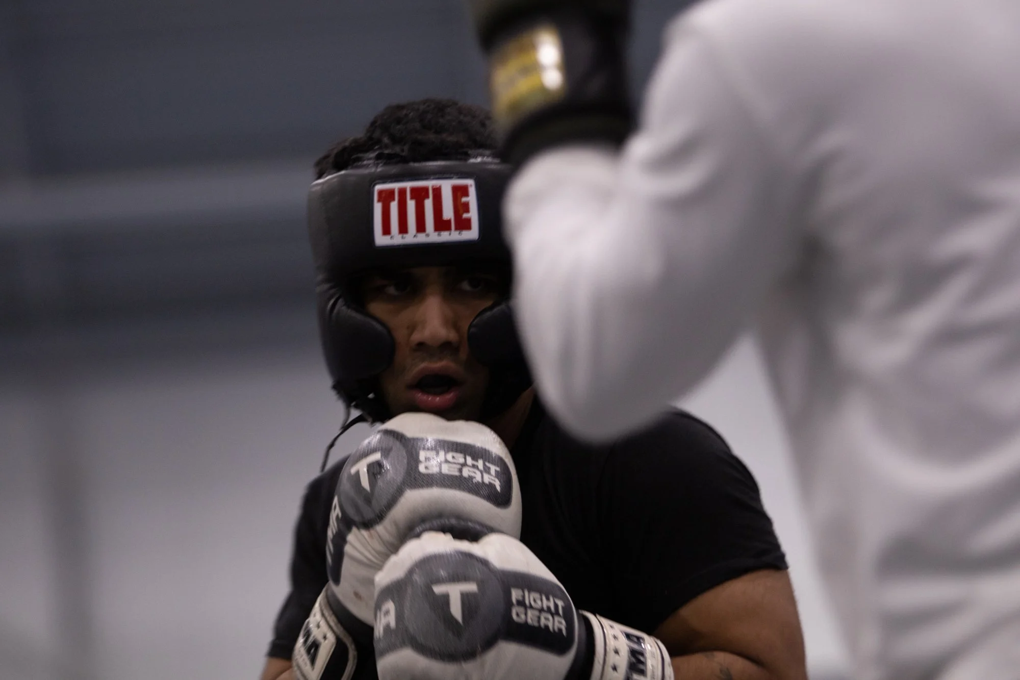 A male boxer wearing a black headgear with 'TITLE' written on it, gloves, and a black and white sports outfit, is practicing in a boxing gym, throwing punches at a punching mitt held by a person in a white shirt.