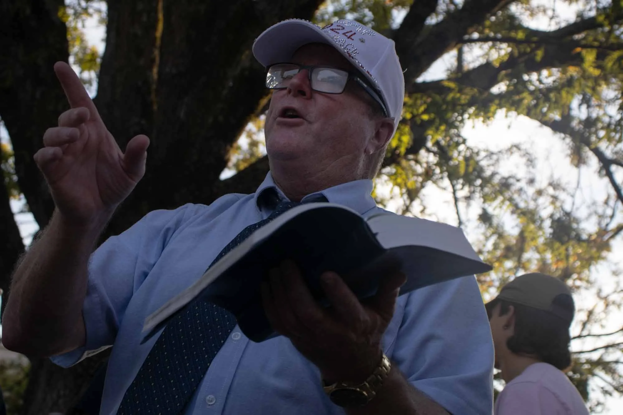 A man wearing glasses, a white cap, and a white shirt with a dark tie is standing outdoors under a large tree with green leaves. The man appears to be speaking or gesturing with his right hand, and the background shows other people and sunlight filte