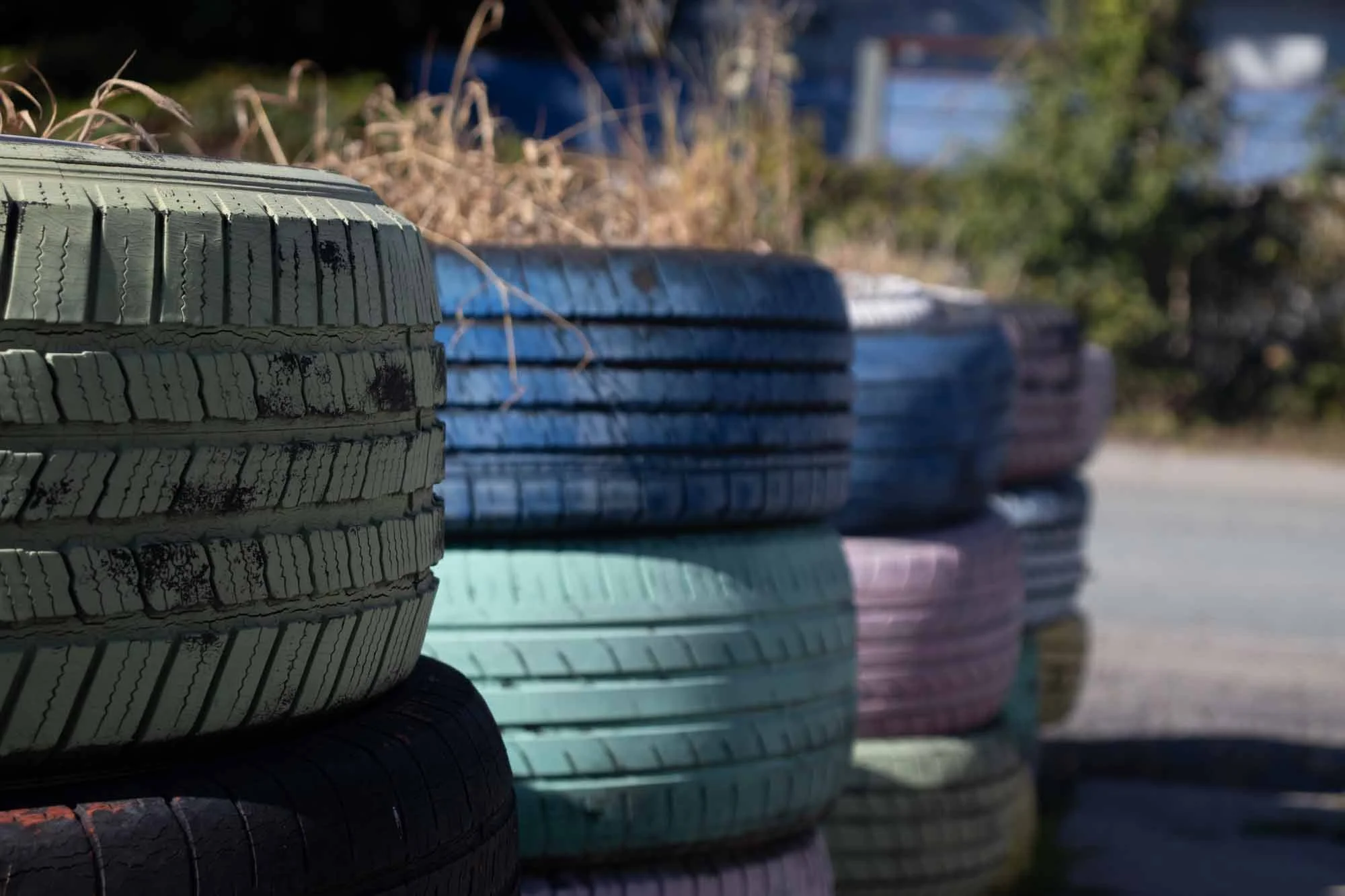 Close-up of stacked tires in various colors, with a background of trees and a blurred blue sky.
