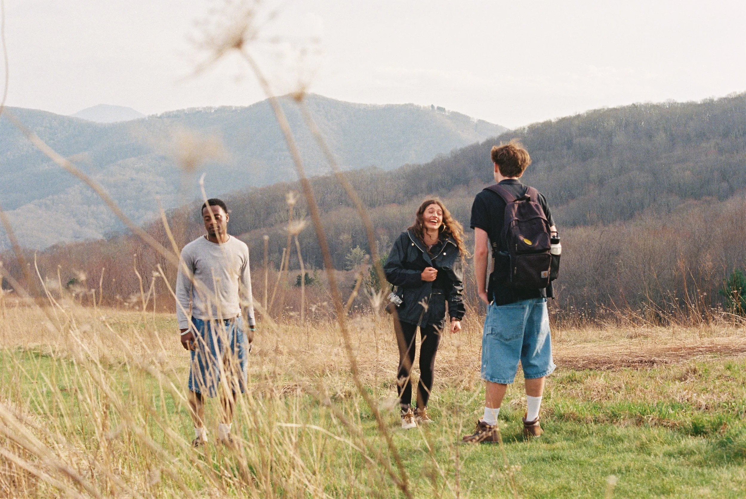 Three young people walking and talking in a grassy field with mountains in the background during the daytime.