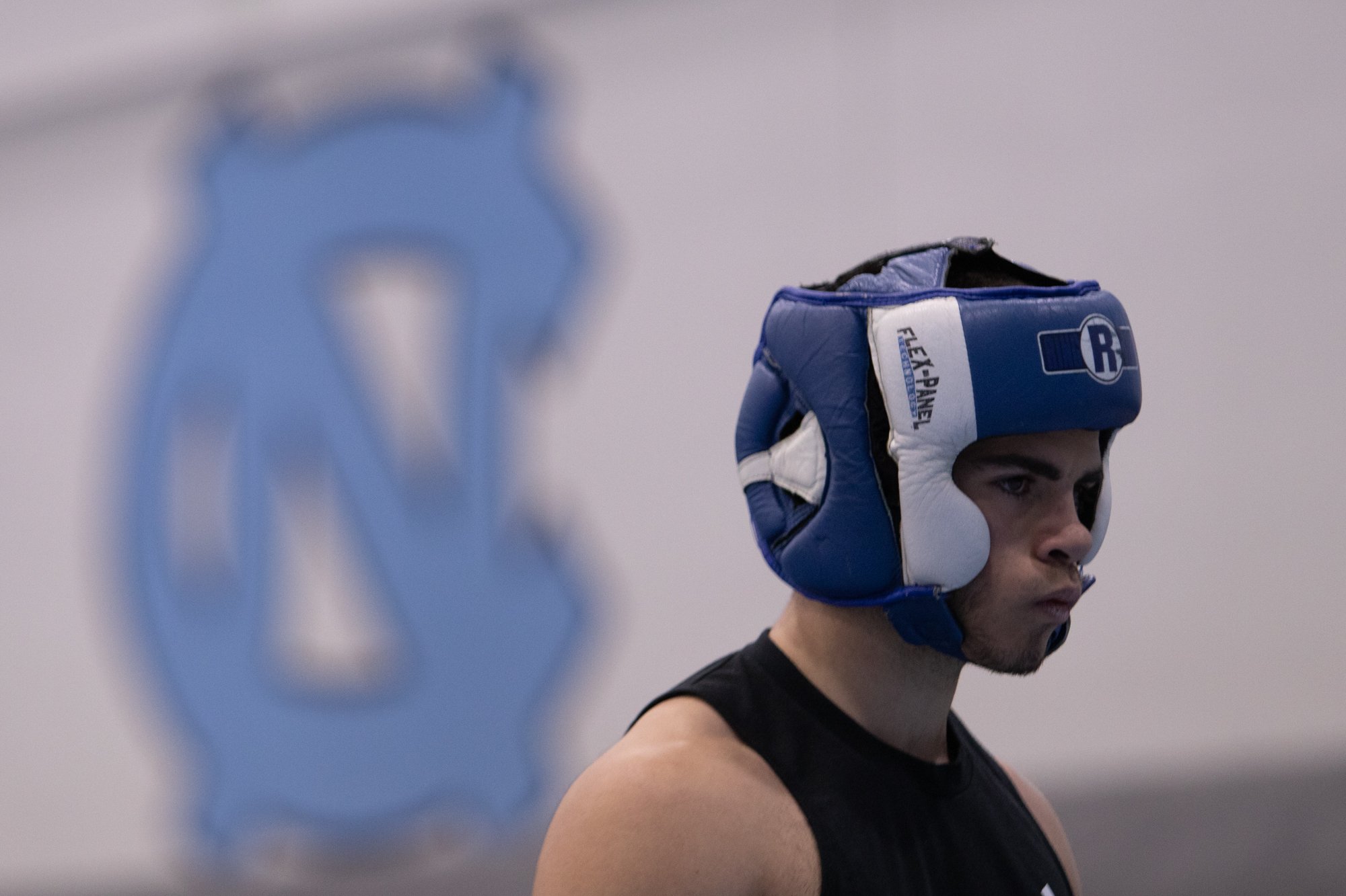A young man wearing a blue boxing helmet, black sleeveless shirt, and athletic build, standing indoors with a serious expression. A blurred blue boxing glove logo is in the background.